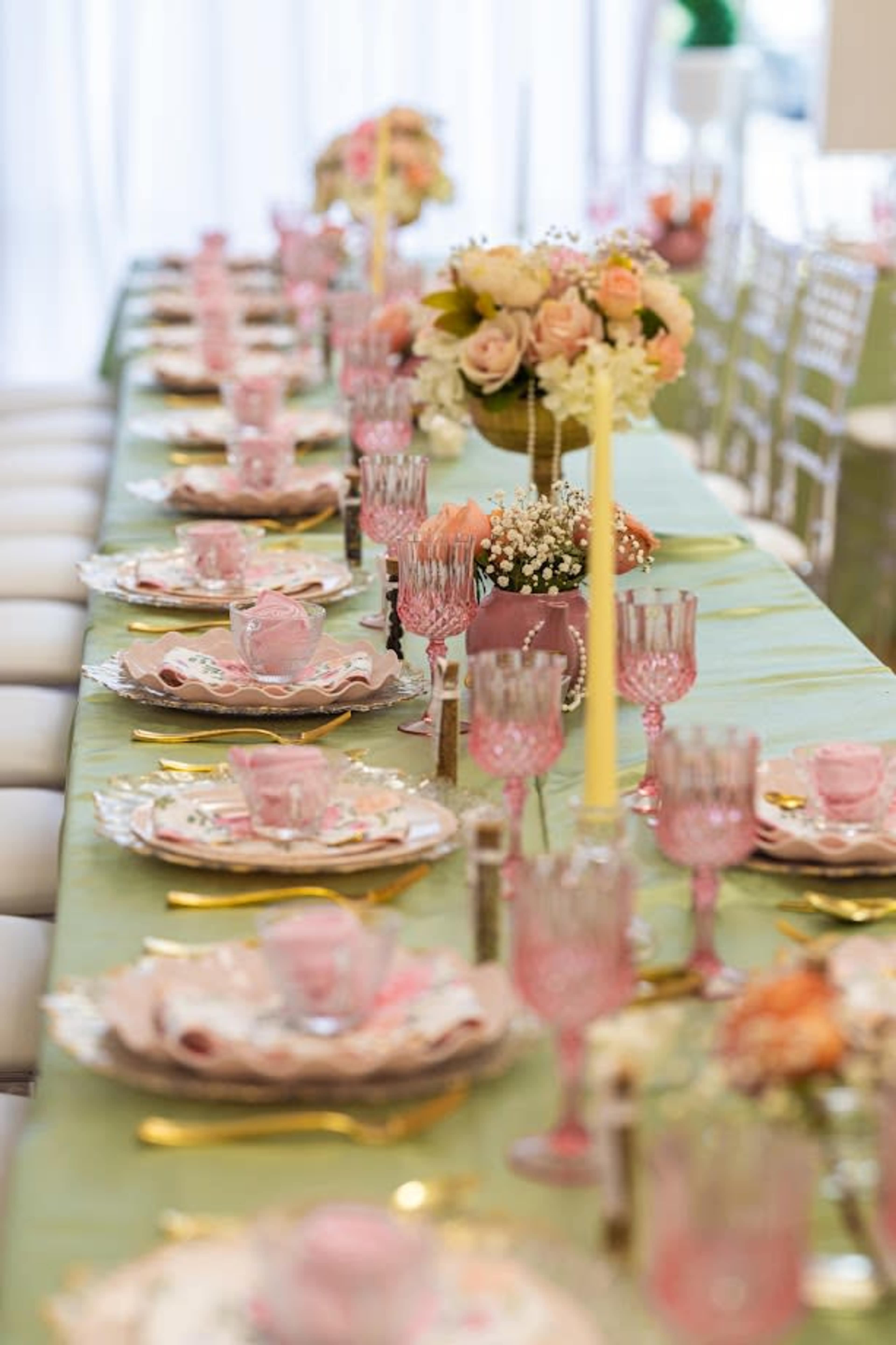 A long dining table is set with elegant pink dishware, glasses, and floral centerpieces, all arranged neatly for a formal gathering.