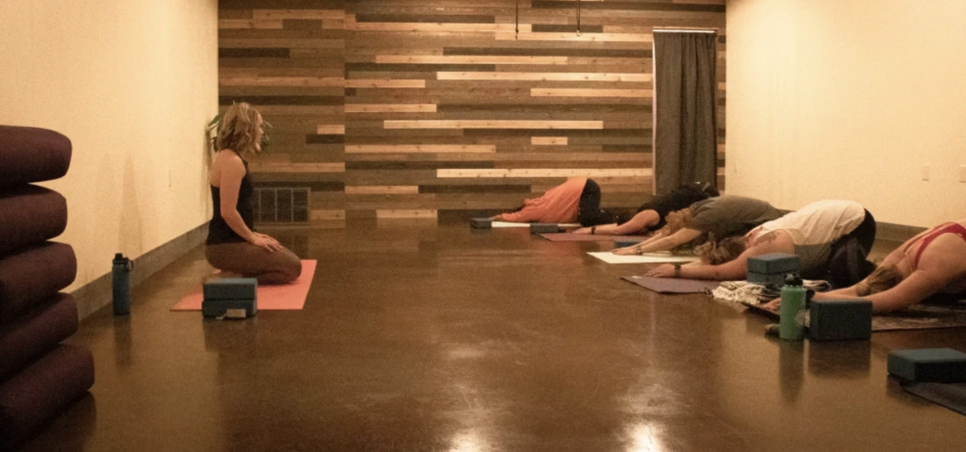 A yoga instructor sits at the front of a dimly lit studio while several participants practice poses on mats in a row.
