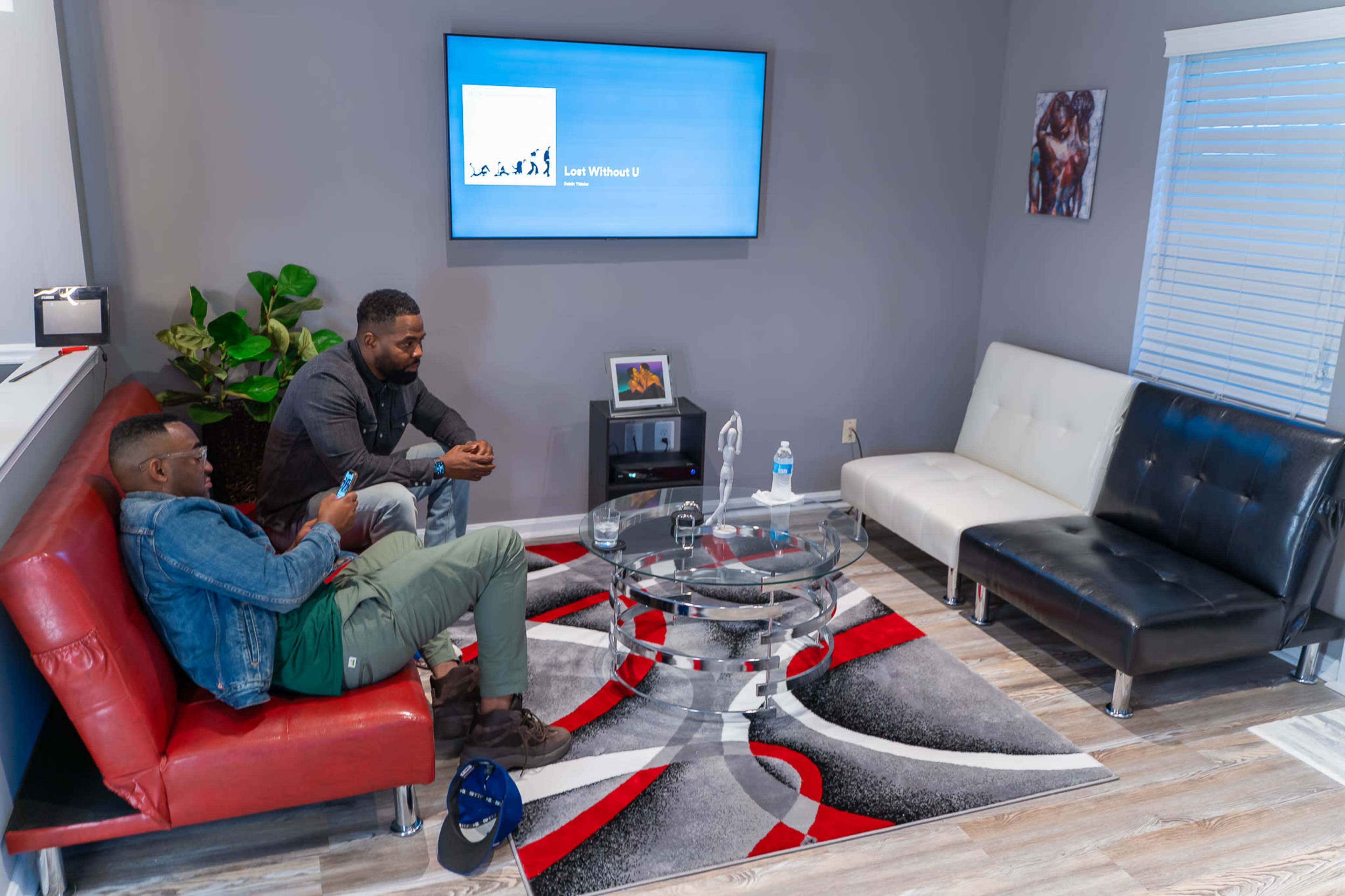 Two men sit in a living room with modern furniture, including a red and black sofa, a glass coffee table, and a television mounted on the wall.