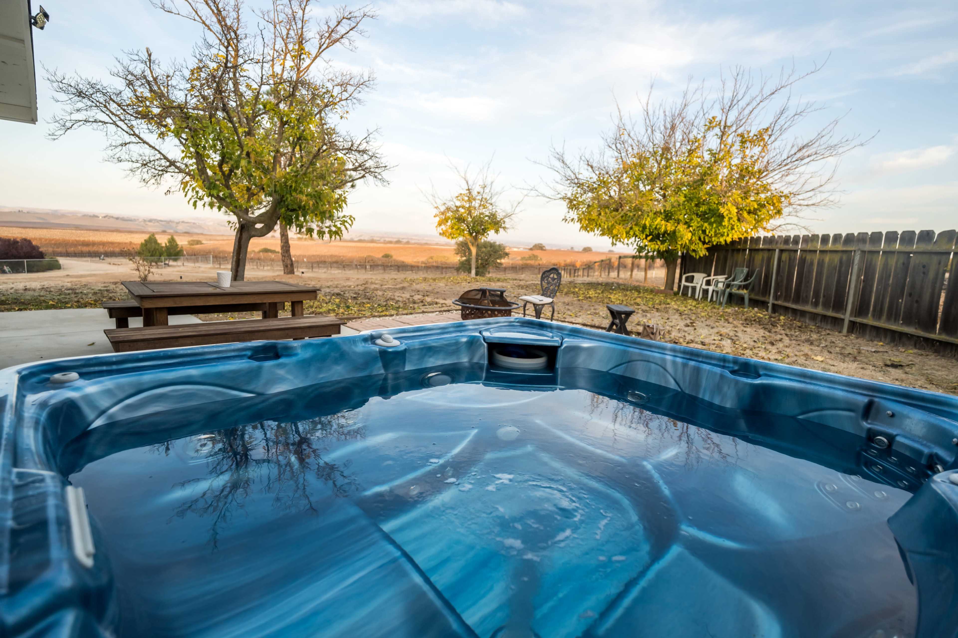 A hot tub is positioned on a patio surrounded by trees and a grassy area under a clear sky.