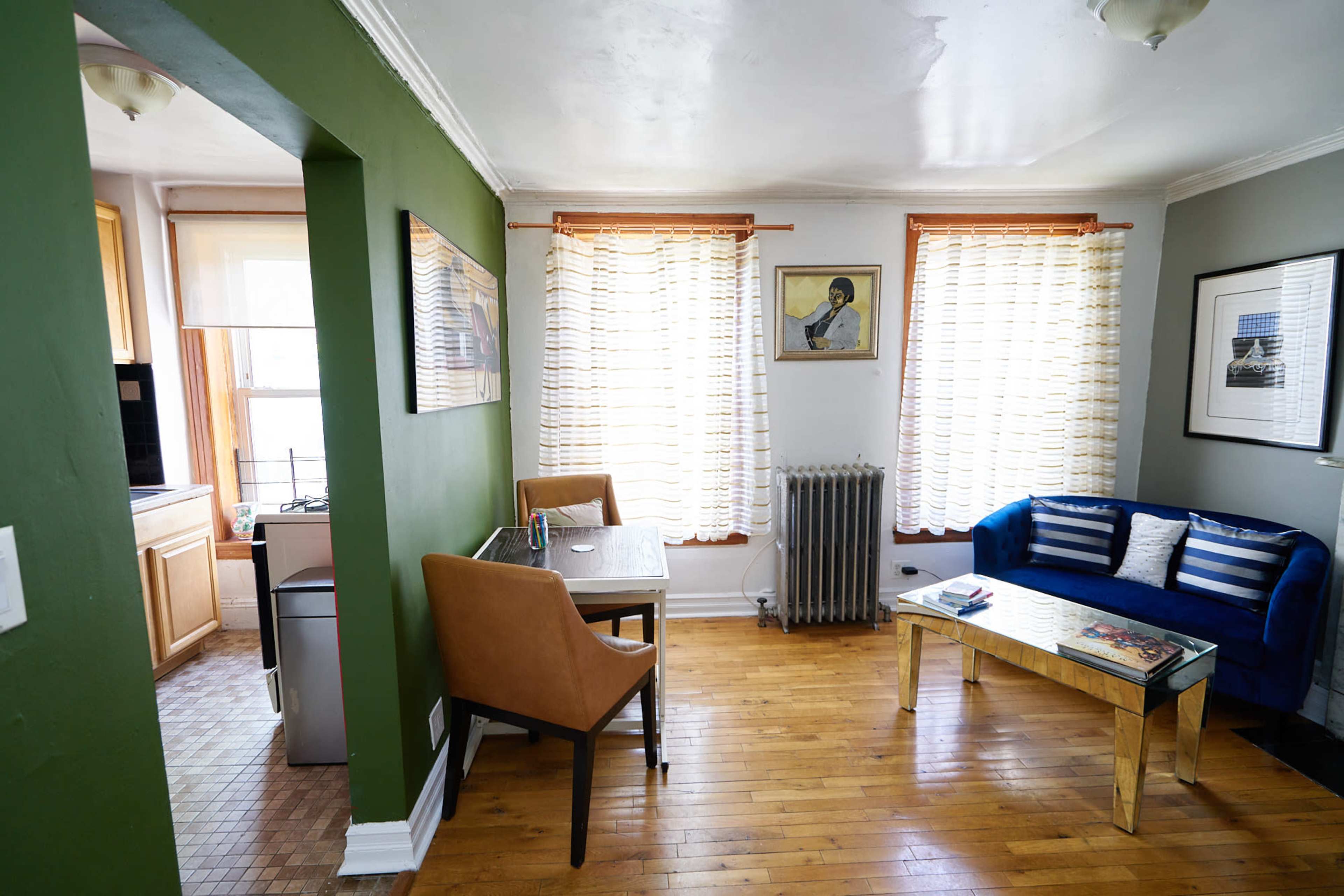 The image shows a well-lit living area with a blue sofa, a wooden dining table, and a vintage radiator, featuring large windows covered with sheer curtains.