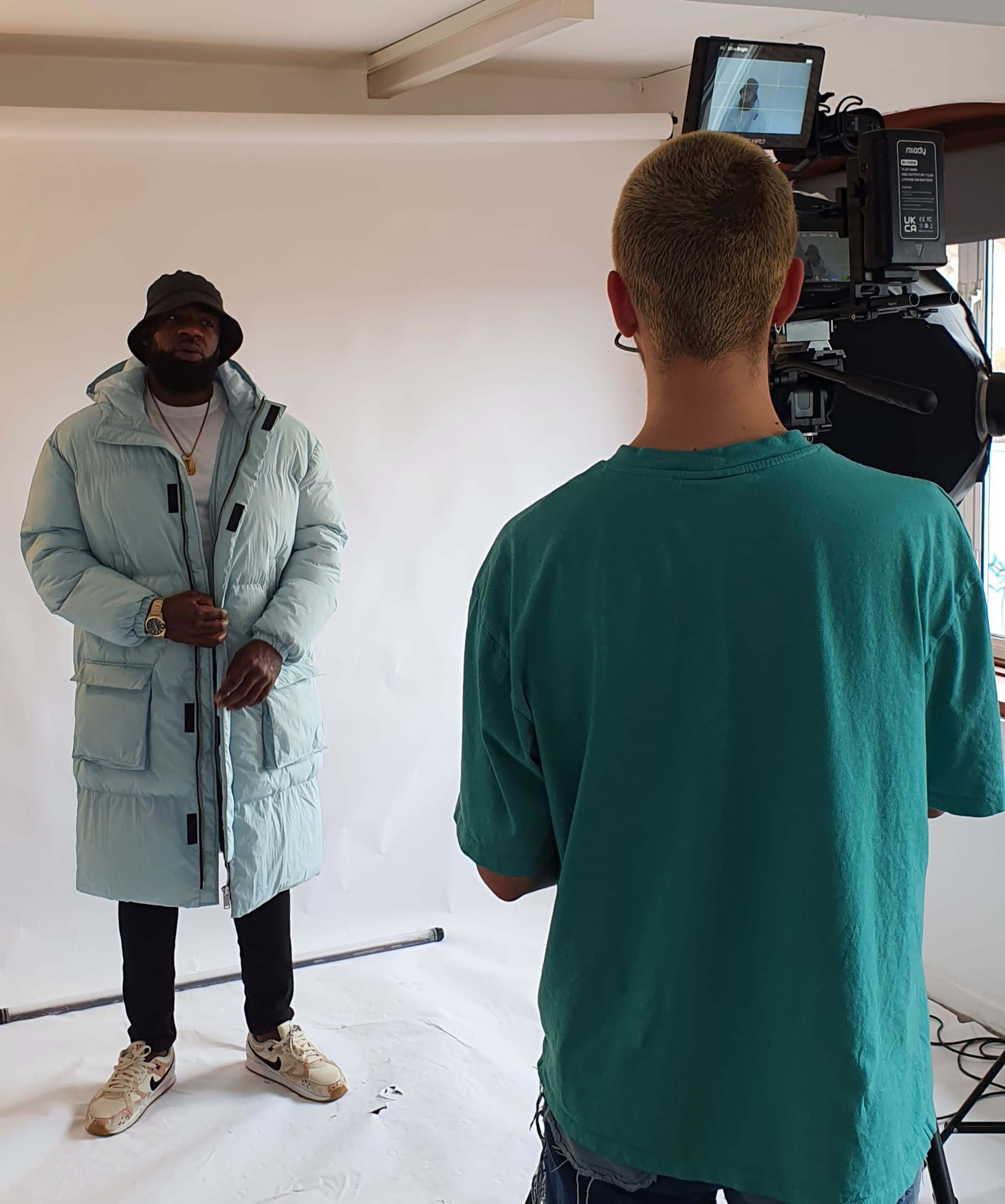 A man in a light blue puffer coat stands in front of a camera, while a crew member operates the equipment from behind.