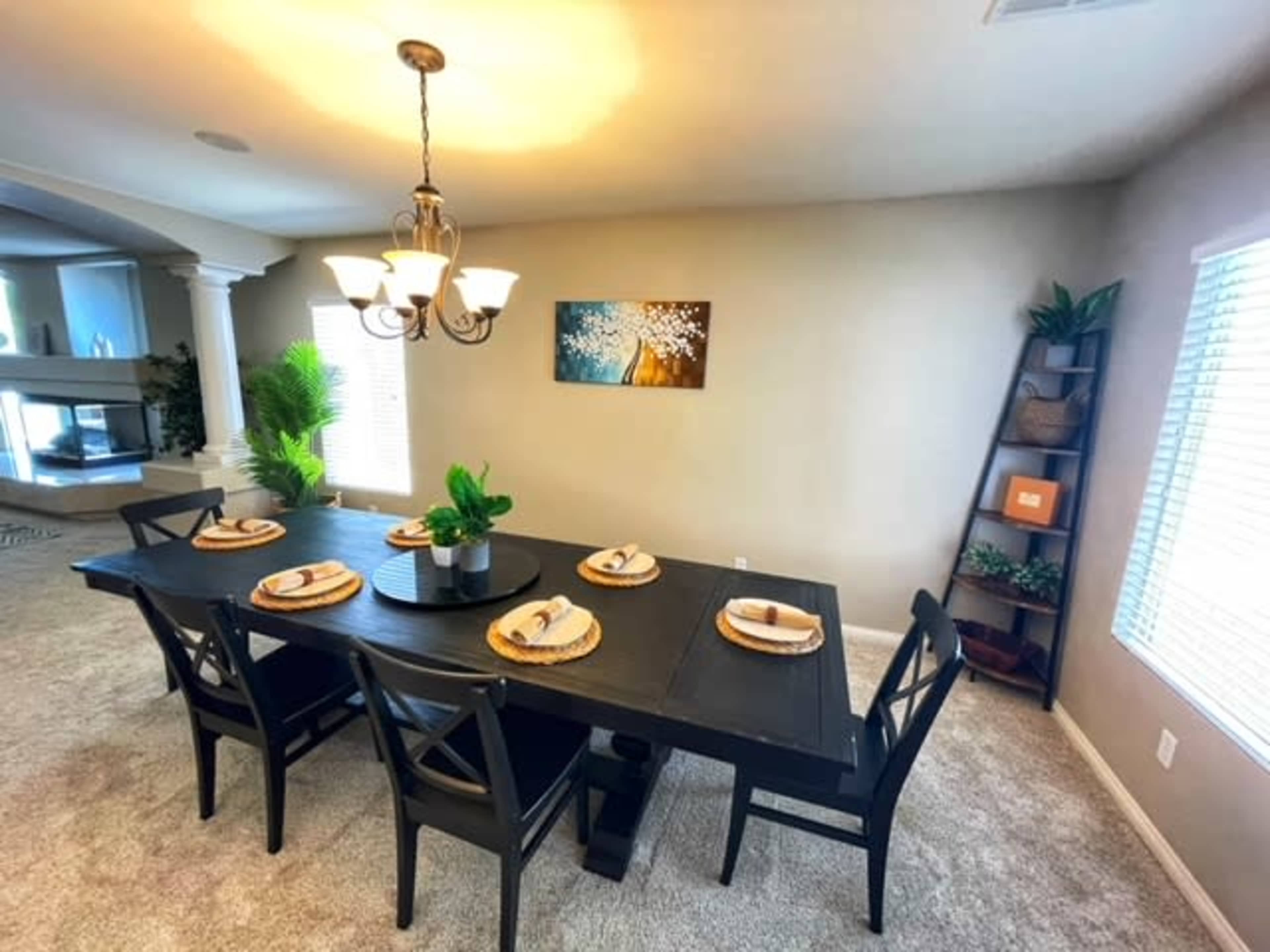 A dining area features a large black table set for six, surrounded by black chairs, with a light fixture above and decorative plants in the background.
