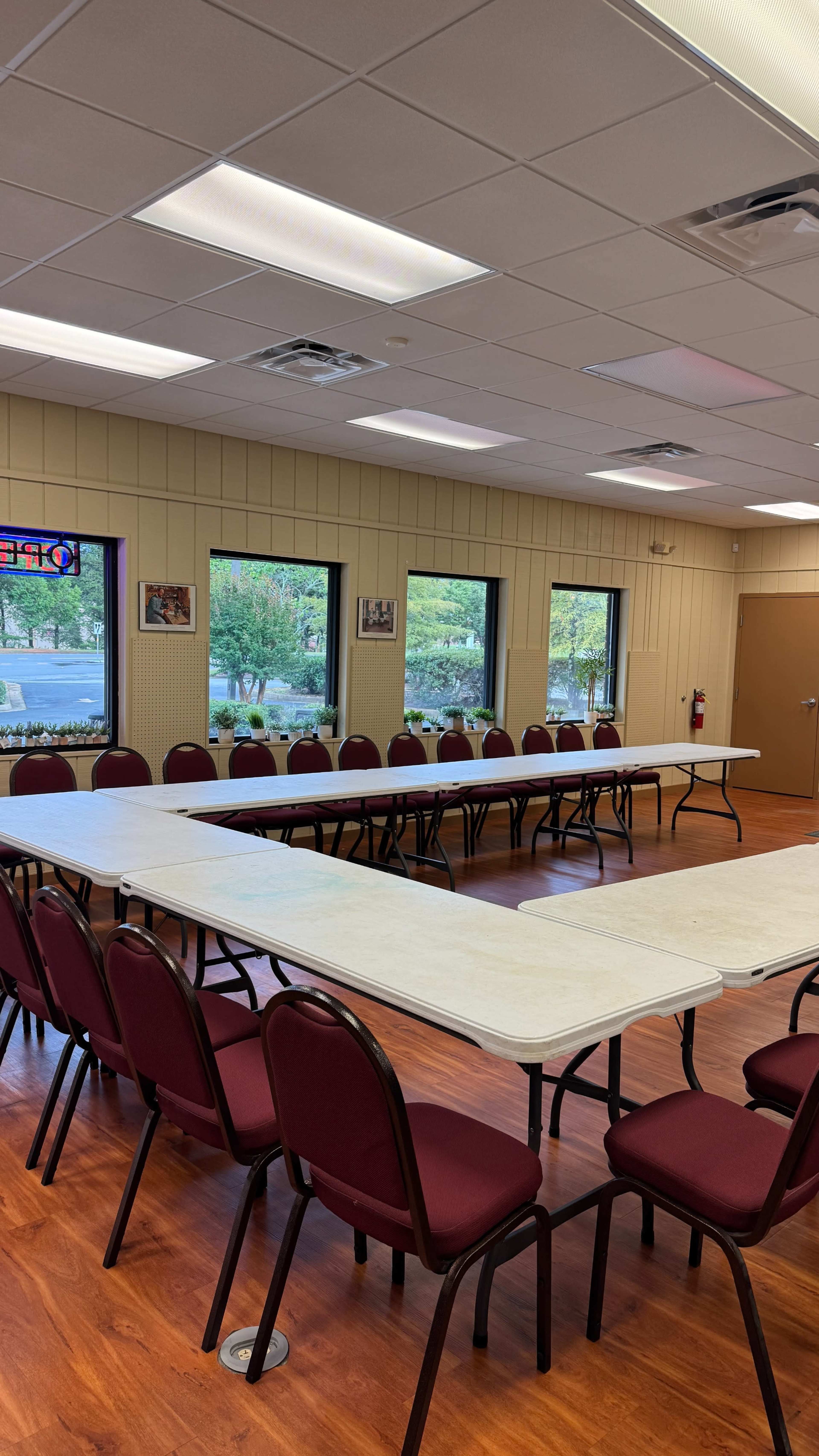 A large meeting room features a long arrangement of white tables and maroon chairs set up neatly, with large windows providing natural light.