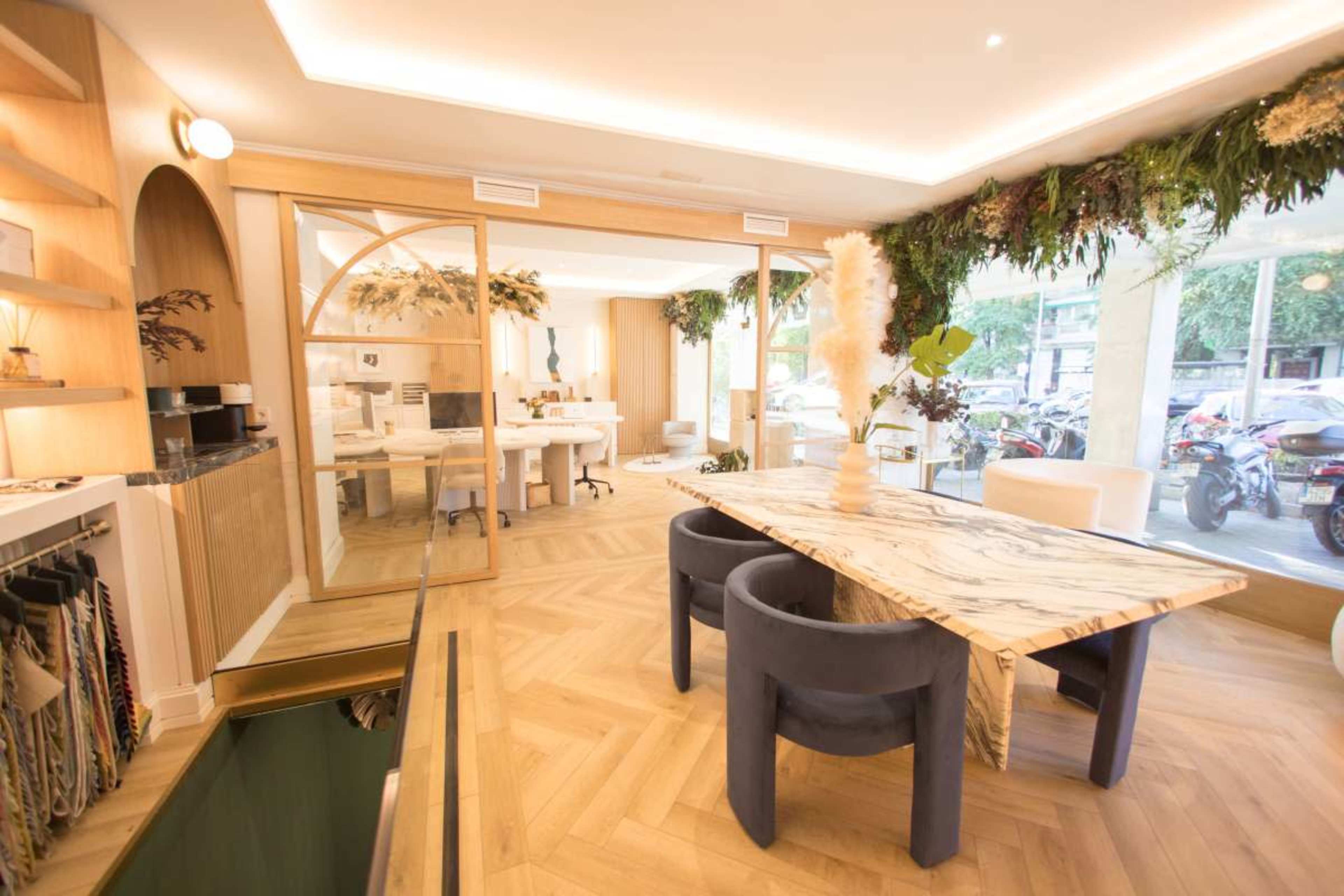 The image shows a modern interior design studio featuring a large marble table surrounded by dark chairs, with greenery and natural light enhancing the space.