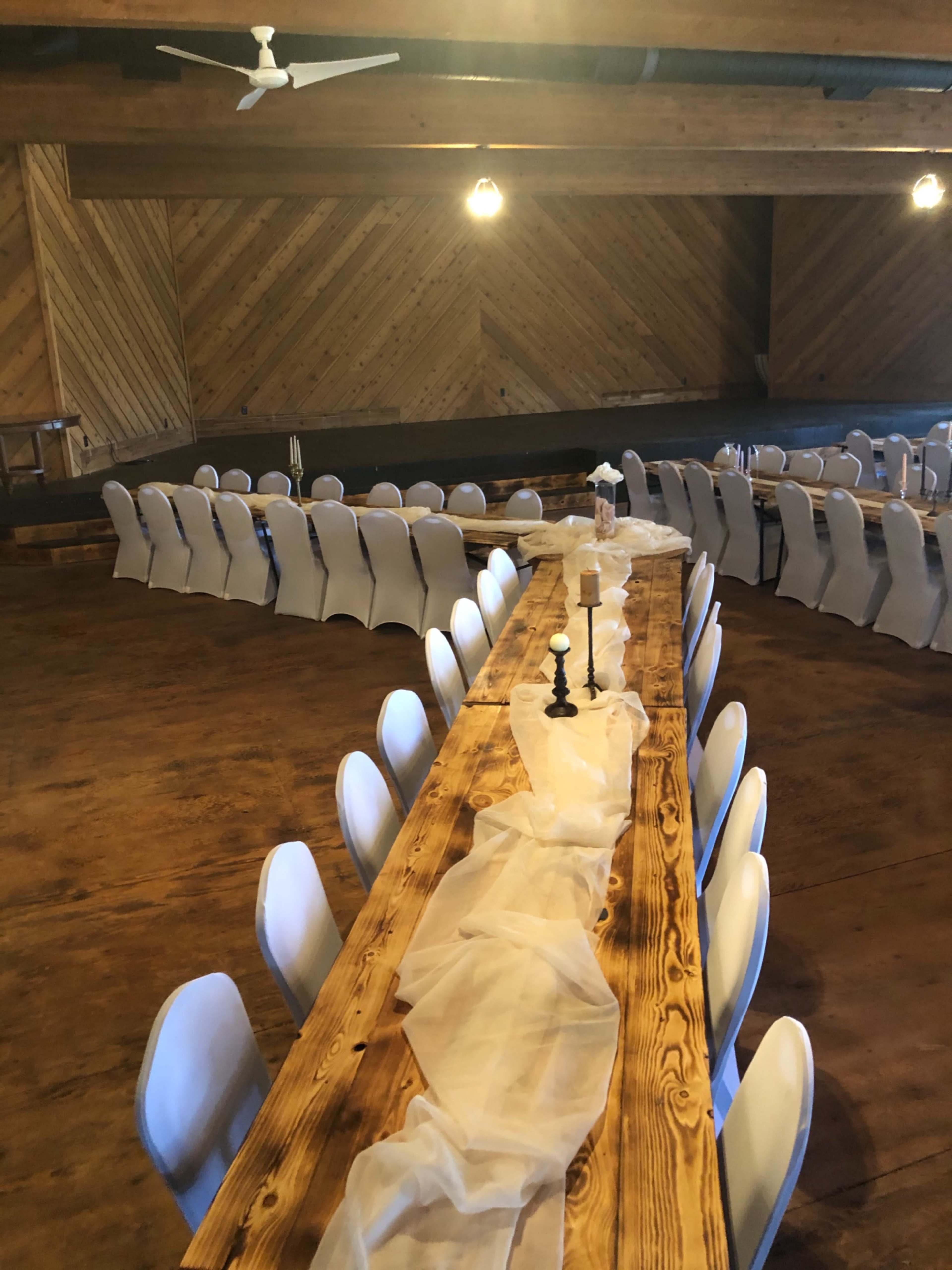 A long wooden table is set with white chairs arranged in a semi-circle, under warm lighting in a wooden-paneled venue.