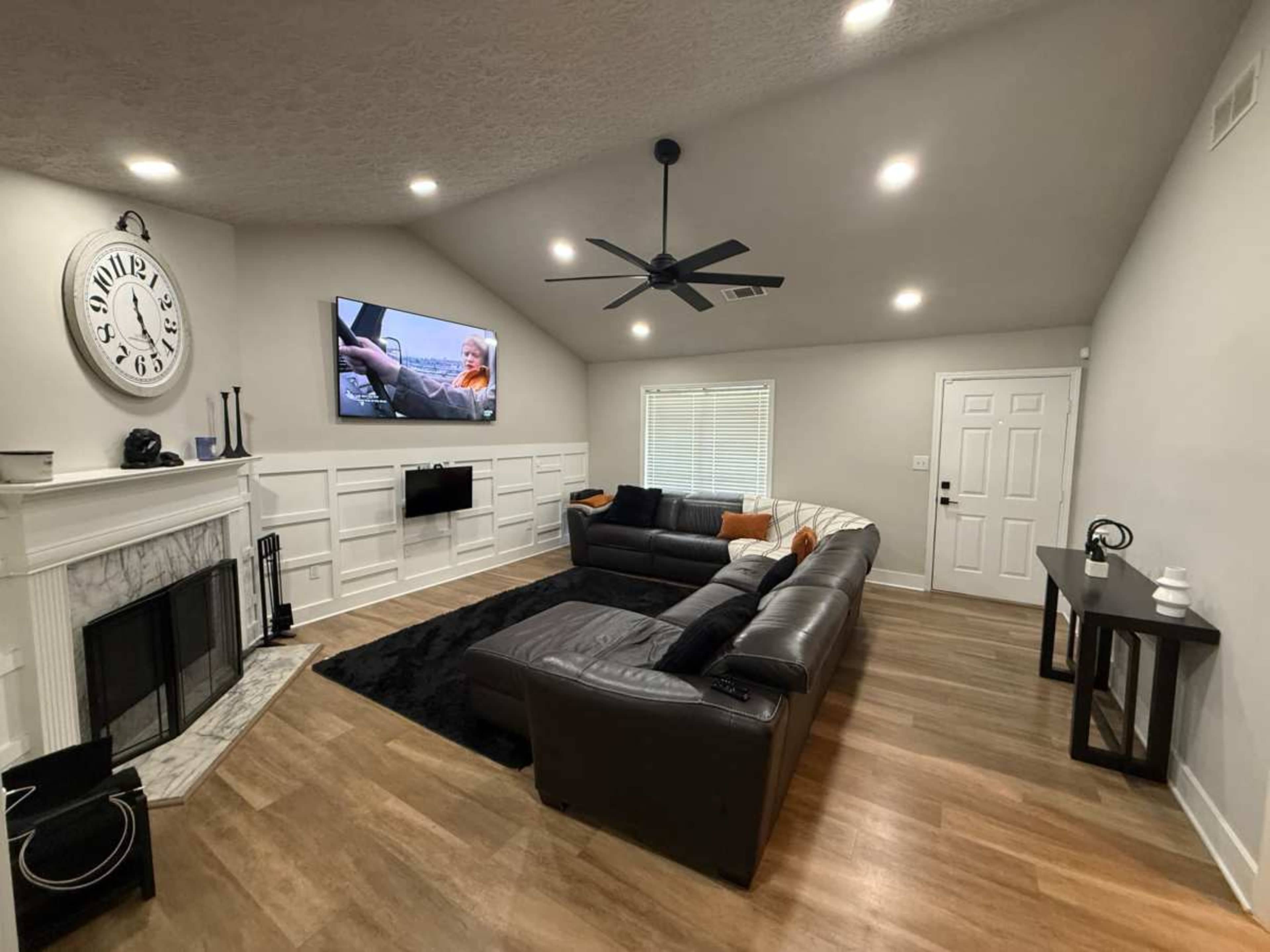 The image shows a modern living room with a dark sectional sofa, a large screen mounted on the wall, and a fireplace beneath a wall clock.