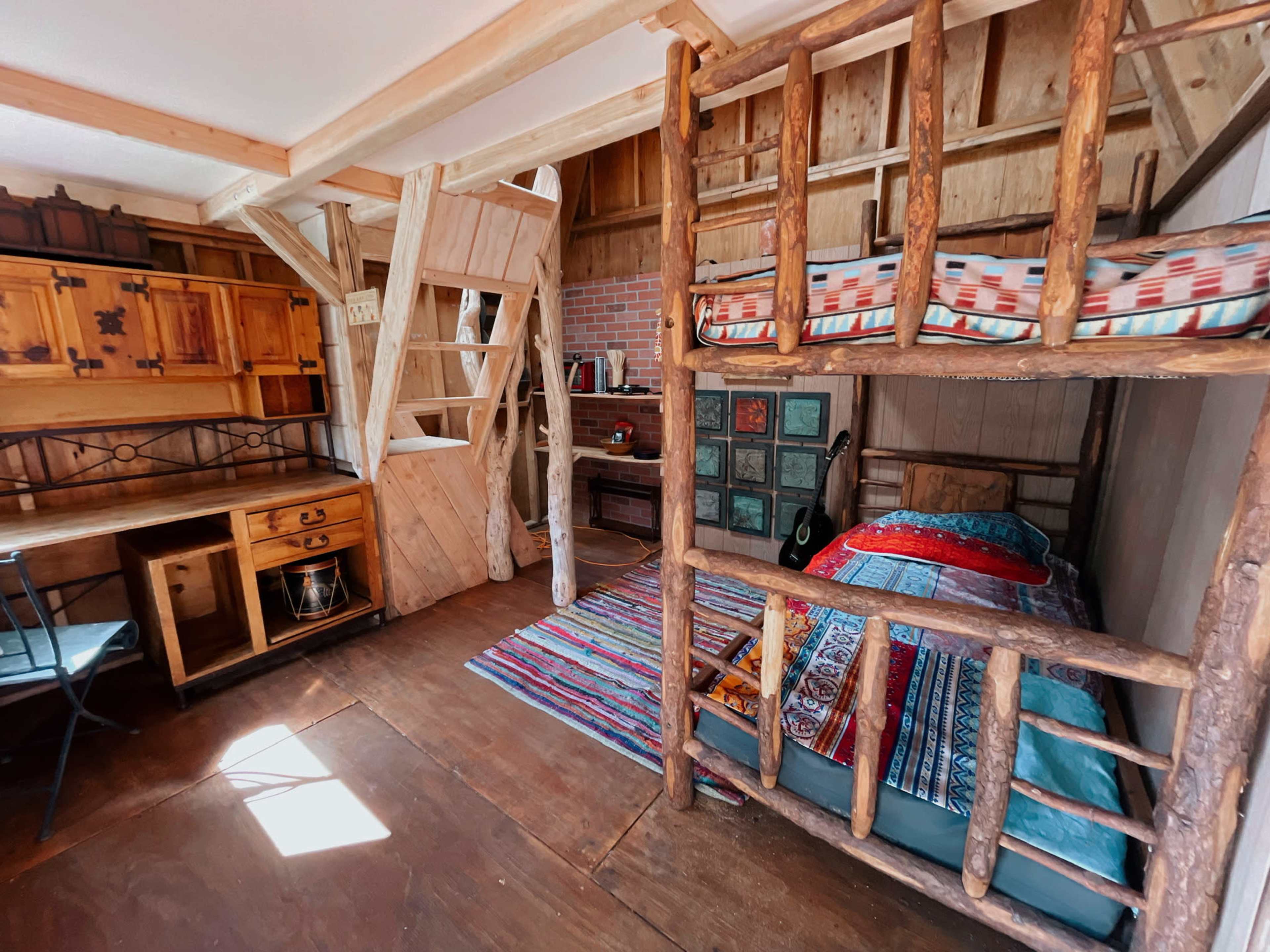 The image shows a cozy wooden cabin interior featuring a bunk bed, a double bed, a rustic desk with a chair, and colorful woven rugs on the floor.