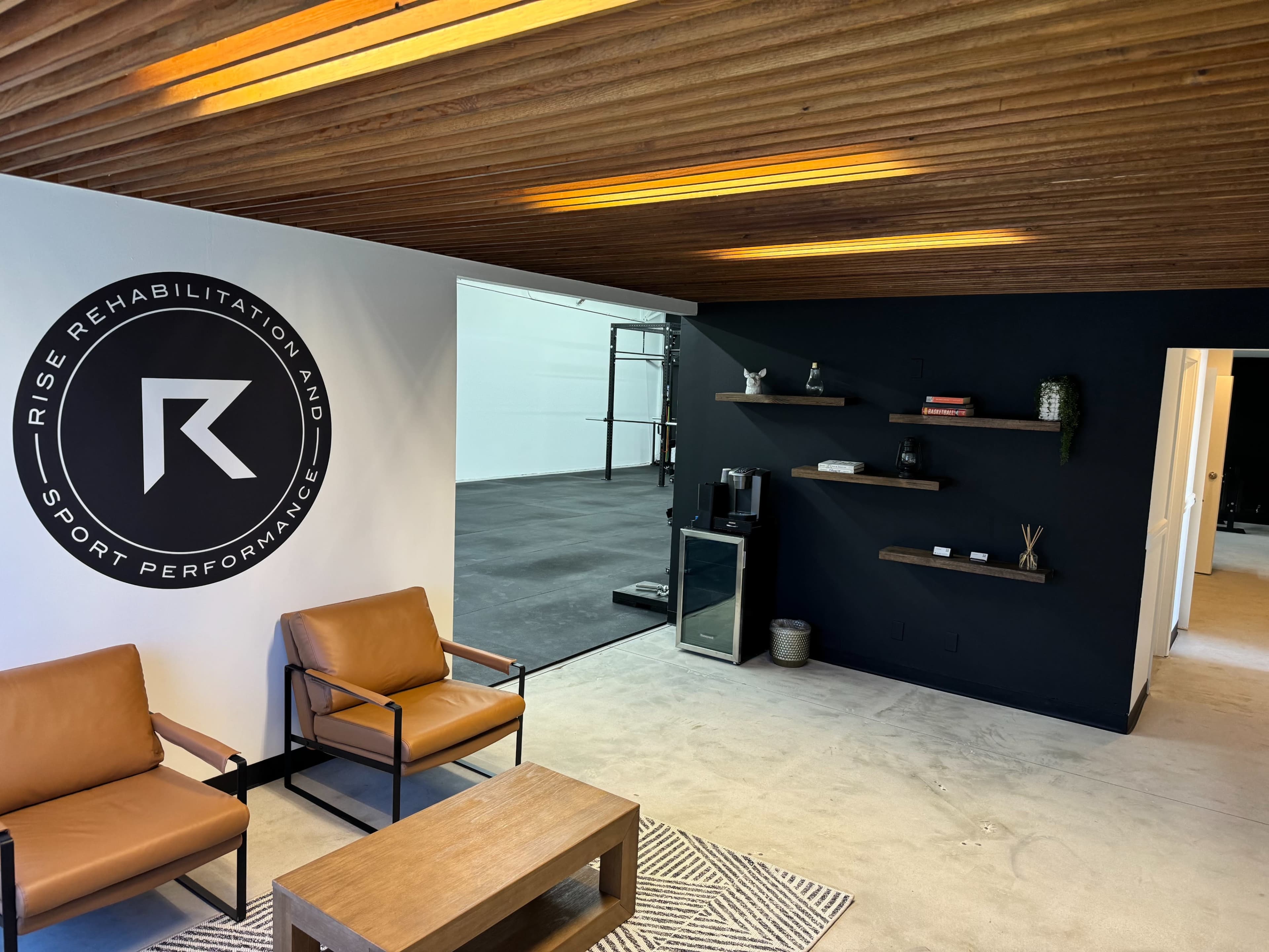 The image shows a modern interior of a rehabilitation and sports performance facility featuring brown leather chairs, a wooden coffee table, and black and white walls with a logo.