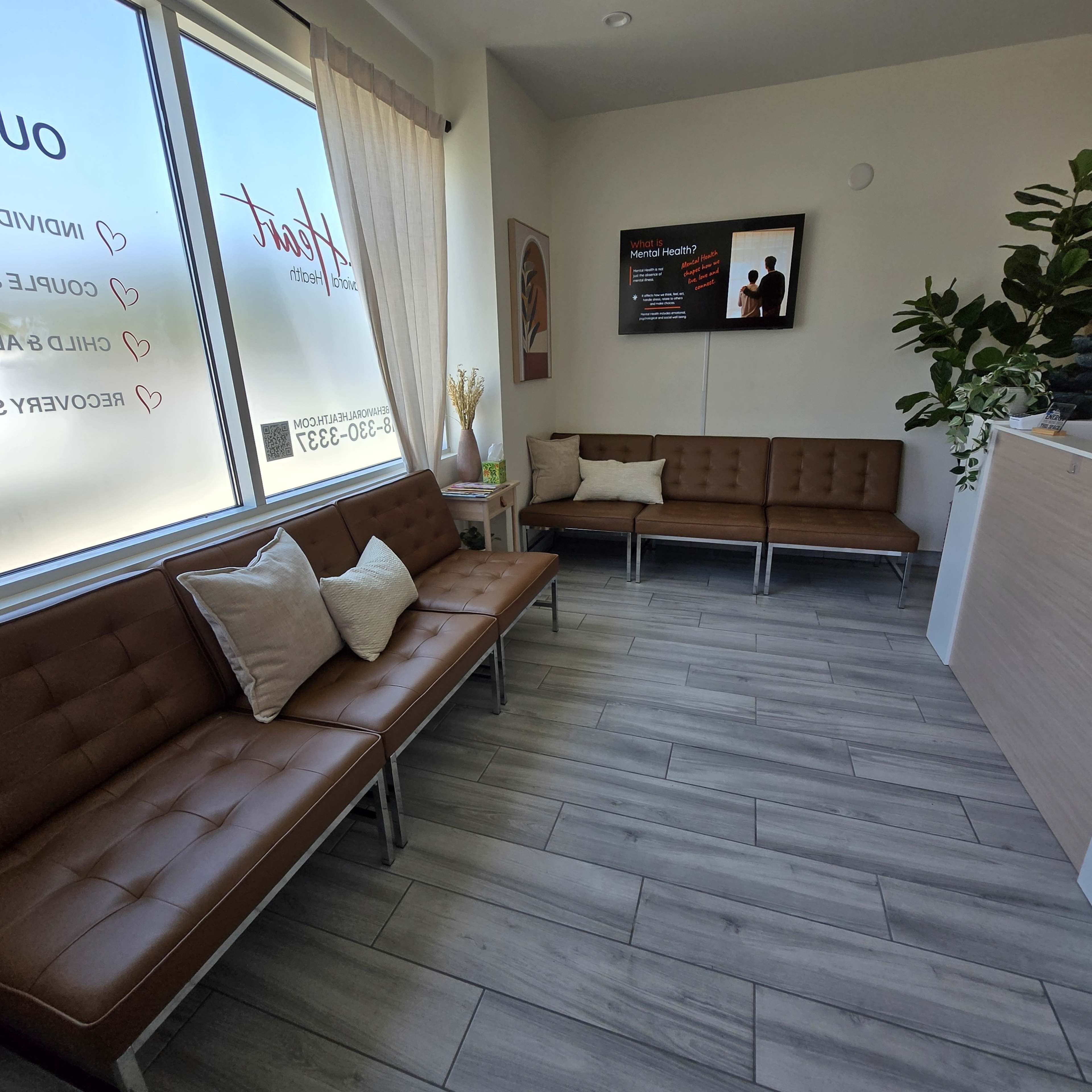 The image shows a modern waiting area with brown tufted couches, a light wooden reception desk, and large windows allowing natural light to enter.