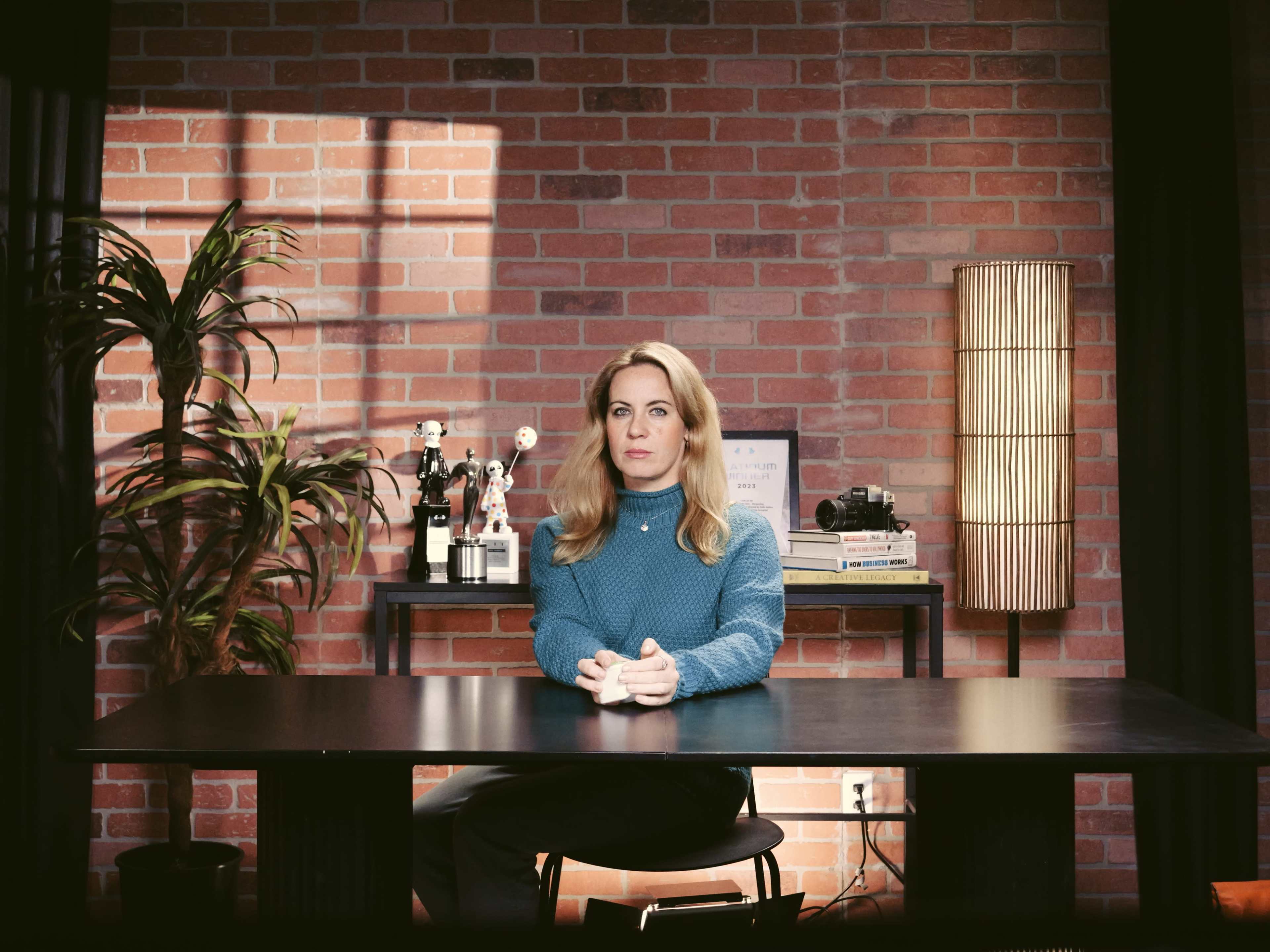 A woman sits at a table in a room with a brick wall, flanked by a plant and a lamp.