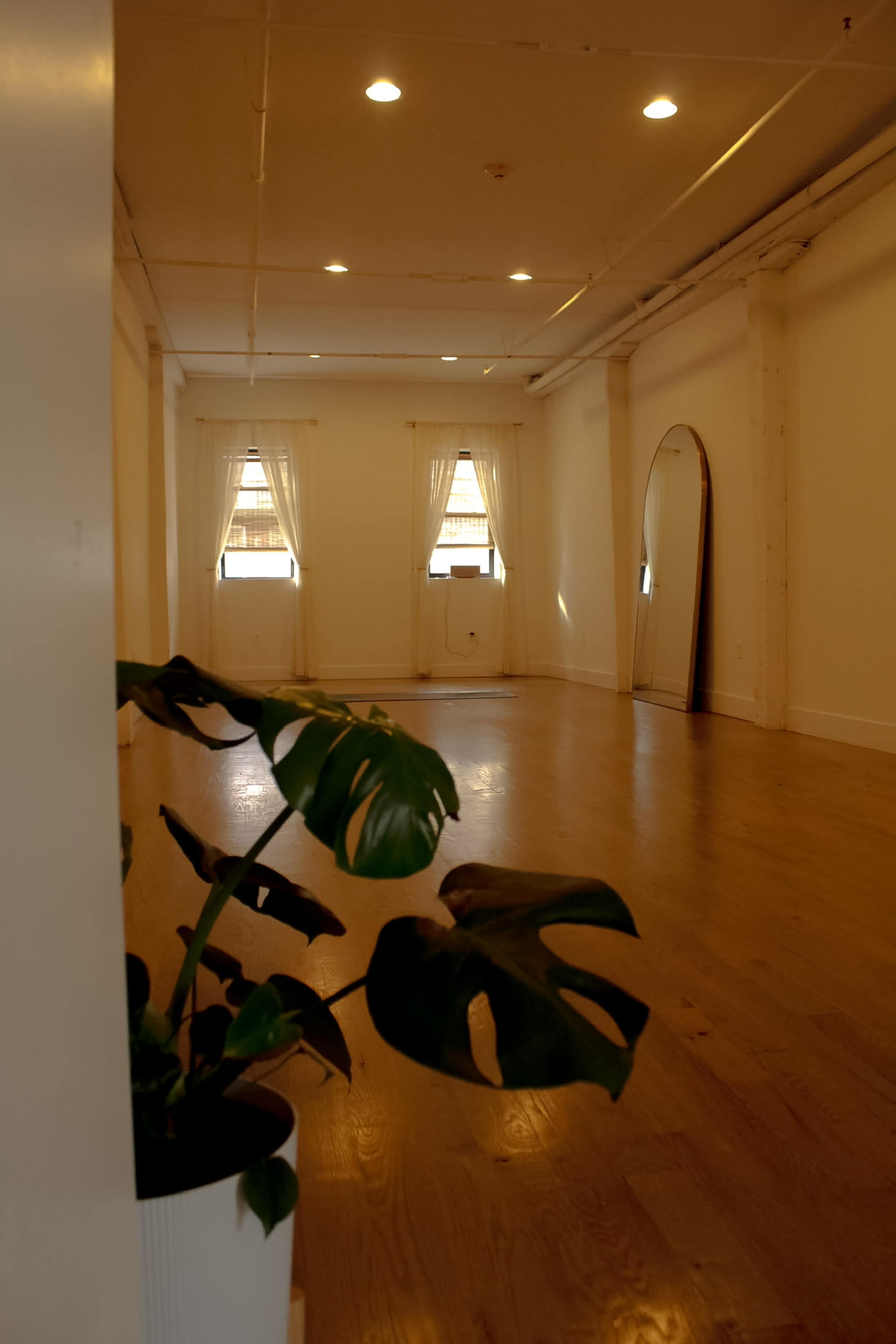 The image shows a bright, minimally furnished room with large windows, hardwood floors, and a standing mirror.