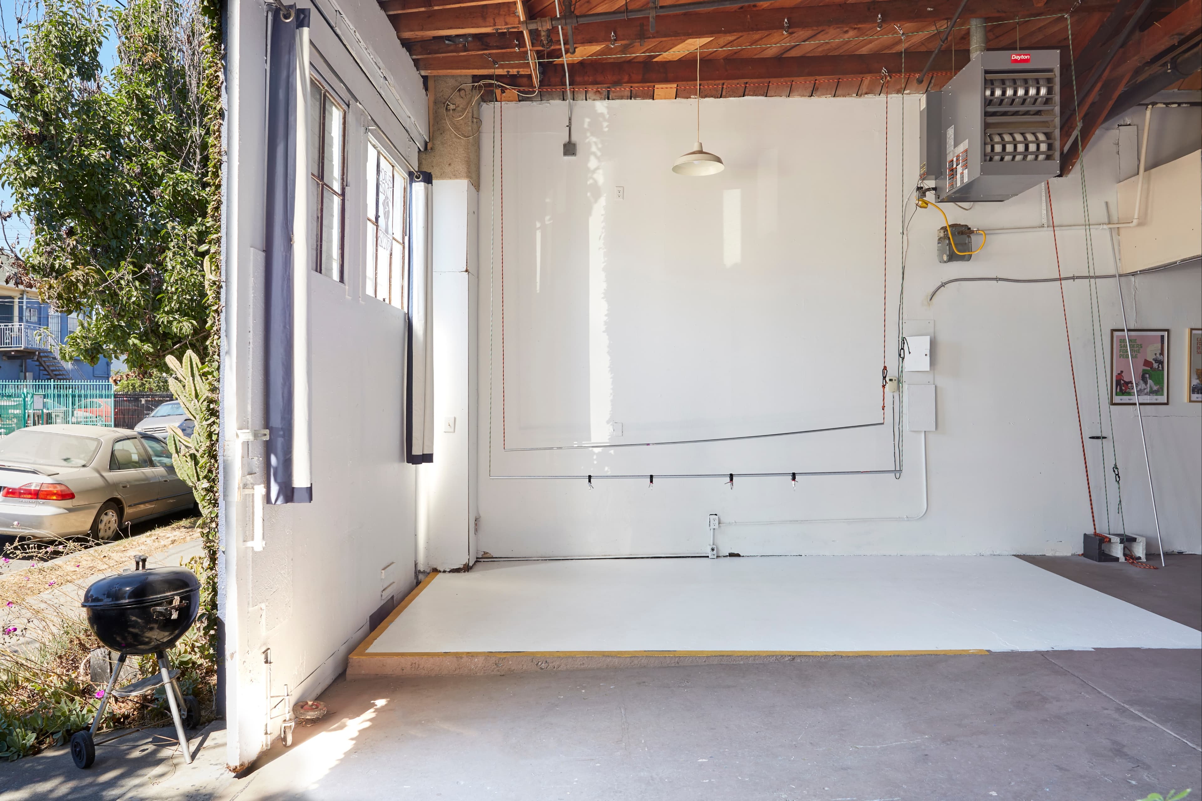An empty room with white walls, a concrete floor, a grill in one corner, and large windows letting in natural light.