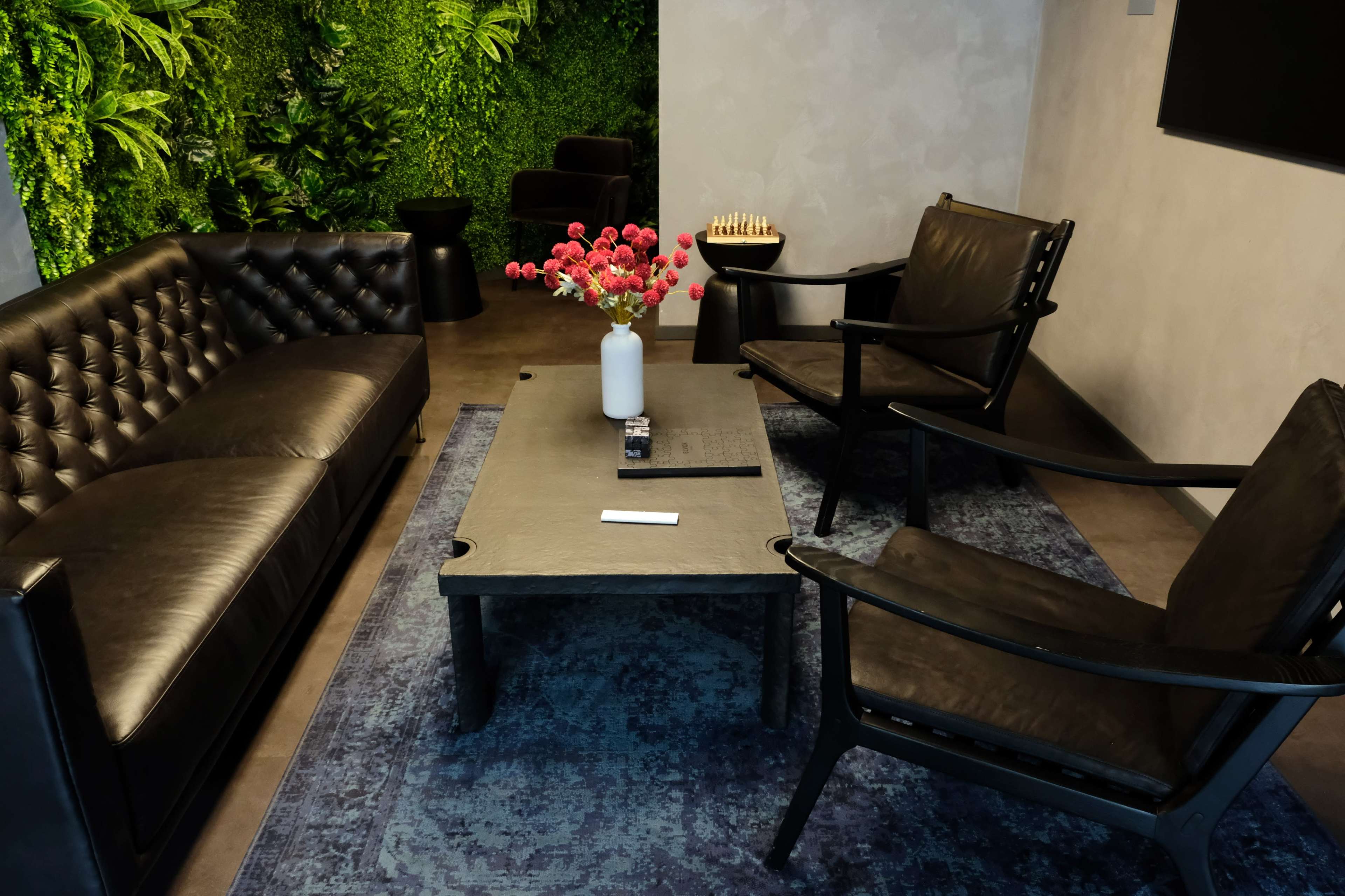 The image shows a modern lounge area with a black leather couch, two black armchairs, a low coffee table, and a wall of greenery in the background.
