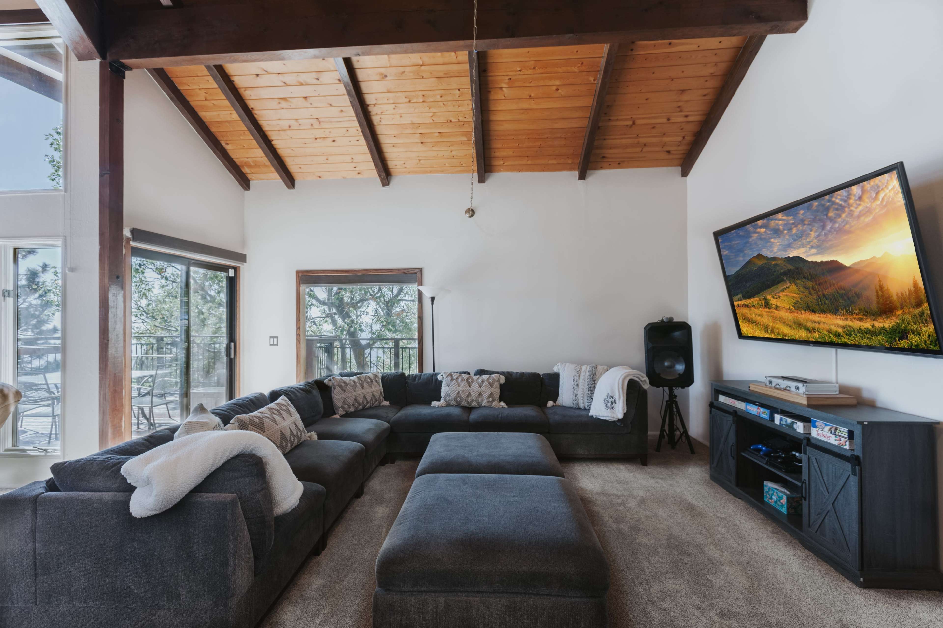 A spacious living room features a large sectional sofa, a mounted television, and wooden beam ceilings with large windows allowing natural light.