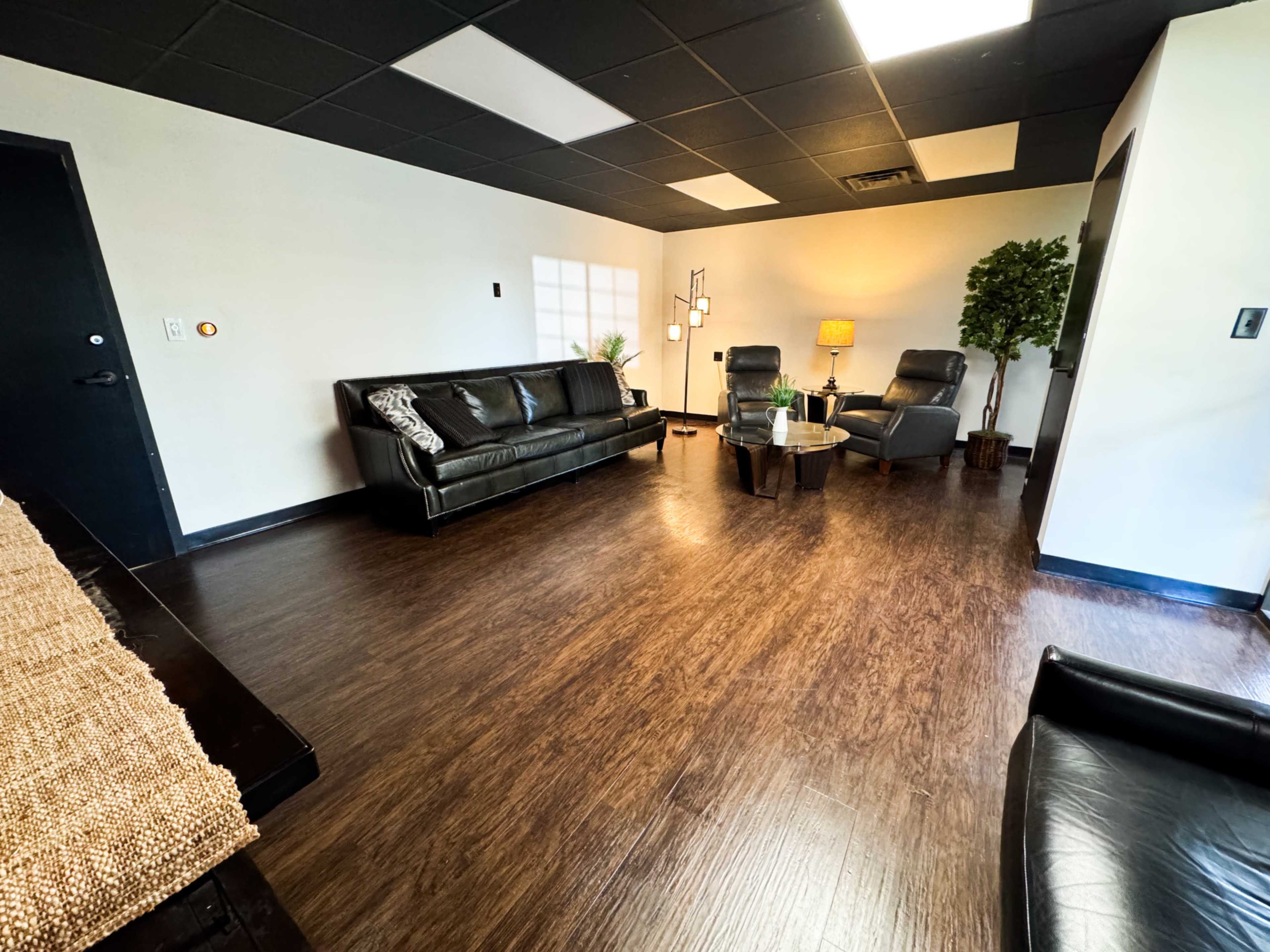 The image shows a modern, minimalist waiting room with black leather sofas, two armchairs, a coffee table, and decorative plants.