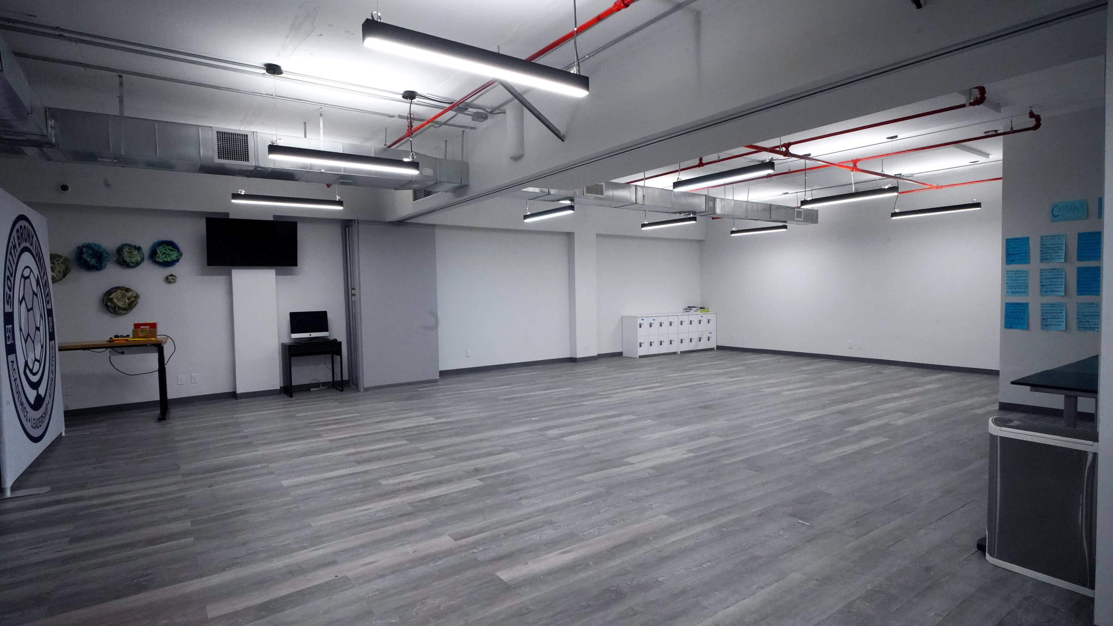 The image shows a spacious, empty room with gray wooden flooring, overhead lights, and minimal furnishings, including a table with a computer and a storage unit along one wall.