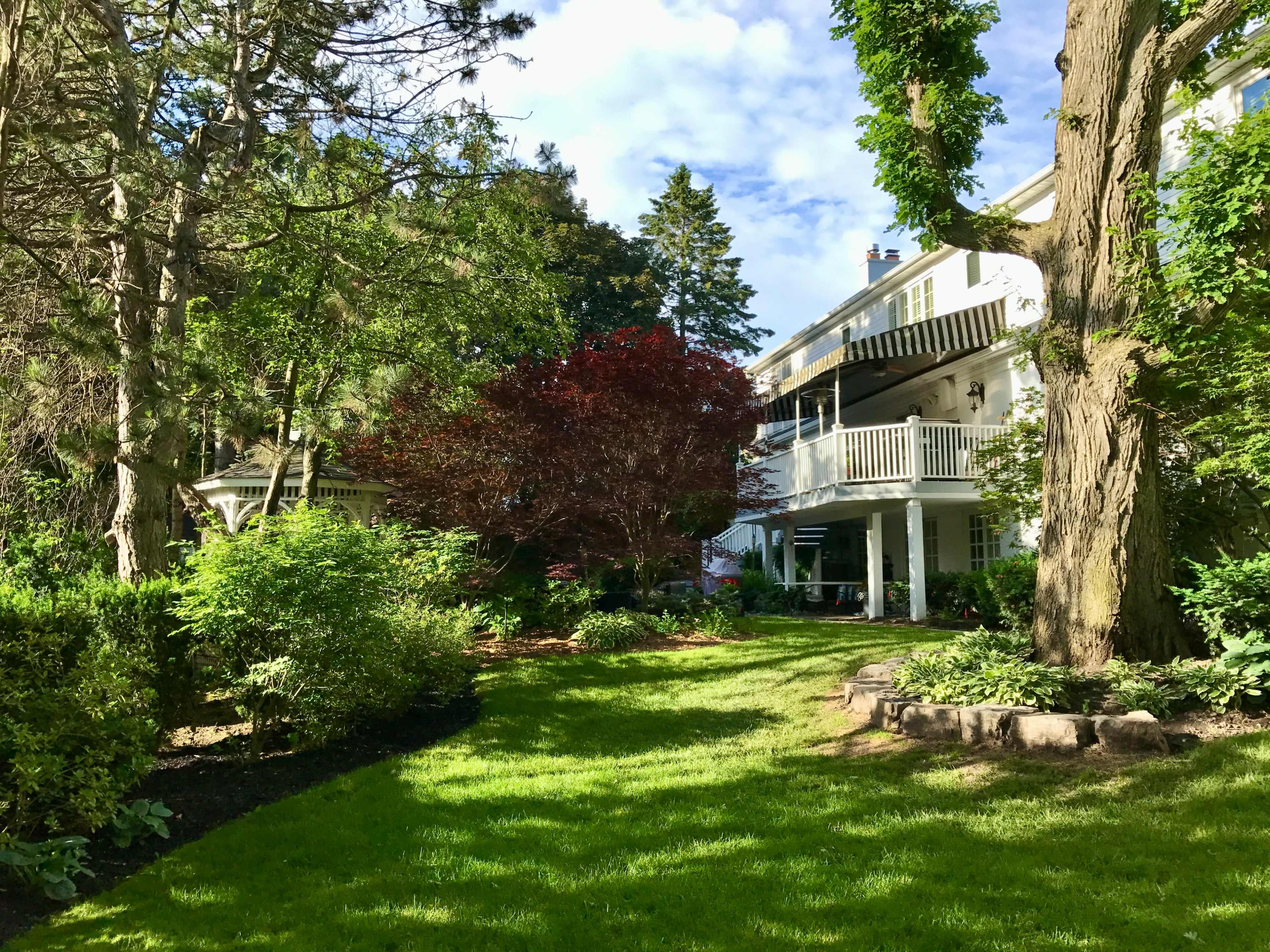 A well-manicured garden features green grass and various trees around a two-story home with a balcony.