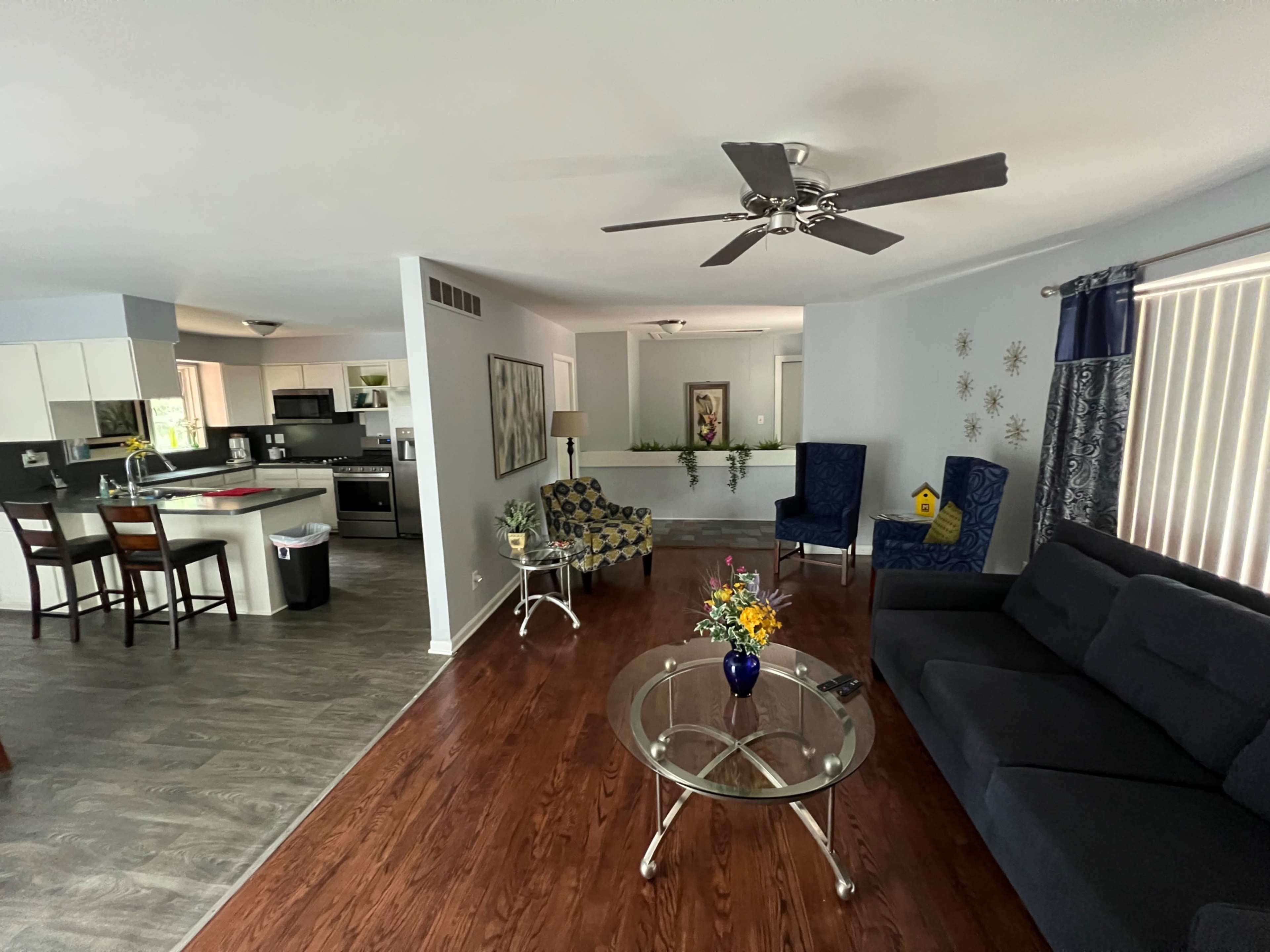 A modern living room area with a gray sofa, a glass coffee table, and two blue chairs, adjacent to a kitchen space with black appliances and a dining area.