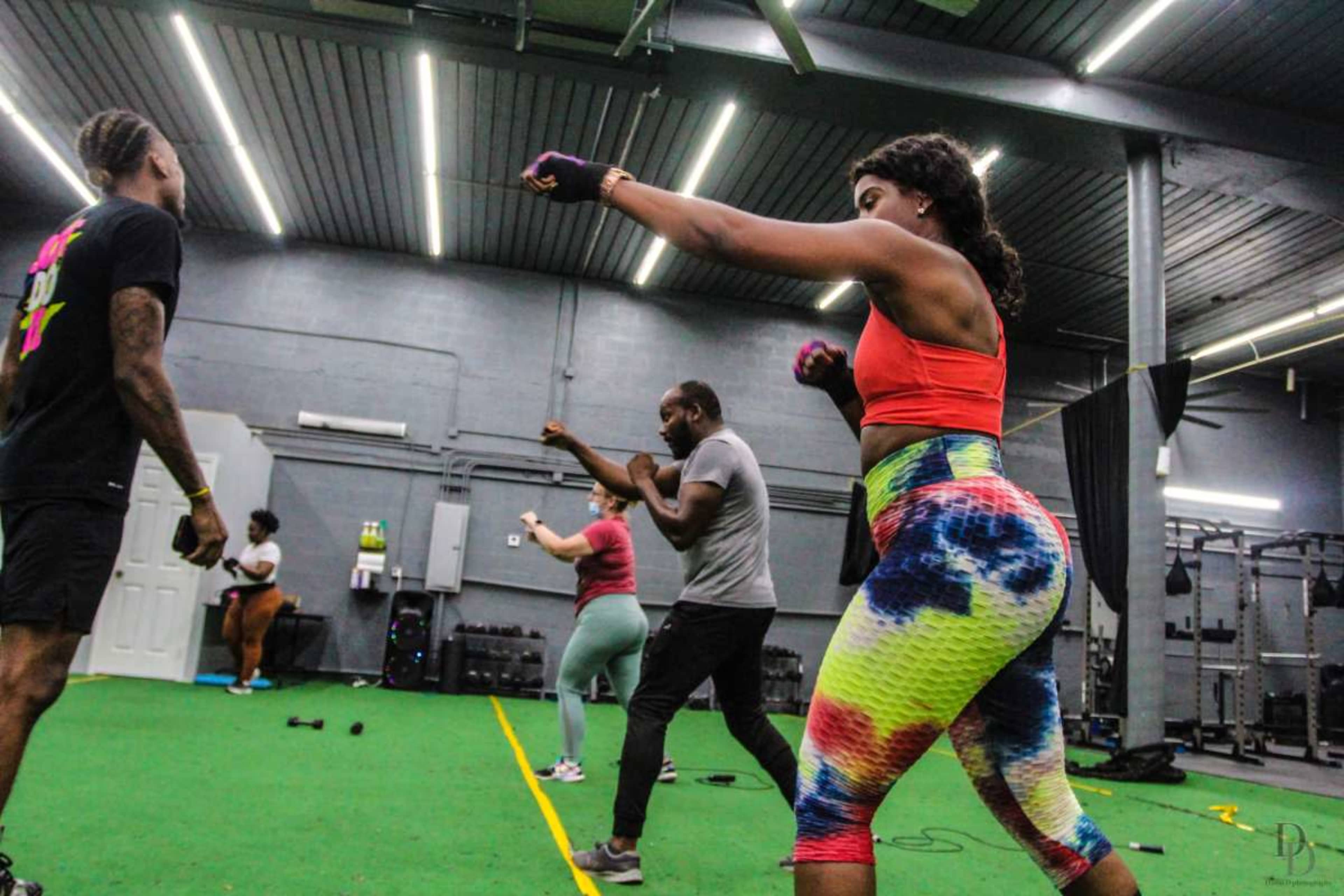A group of people is engaged in a workout session in a gym with green turf flooring, featuring various fitness activities and equipment.