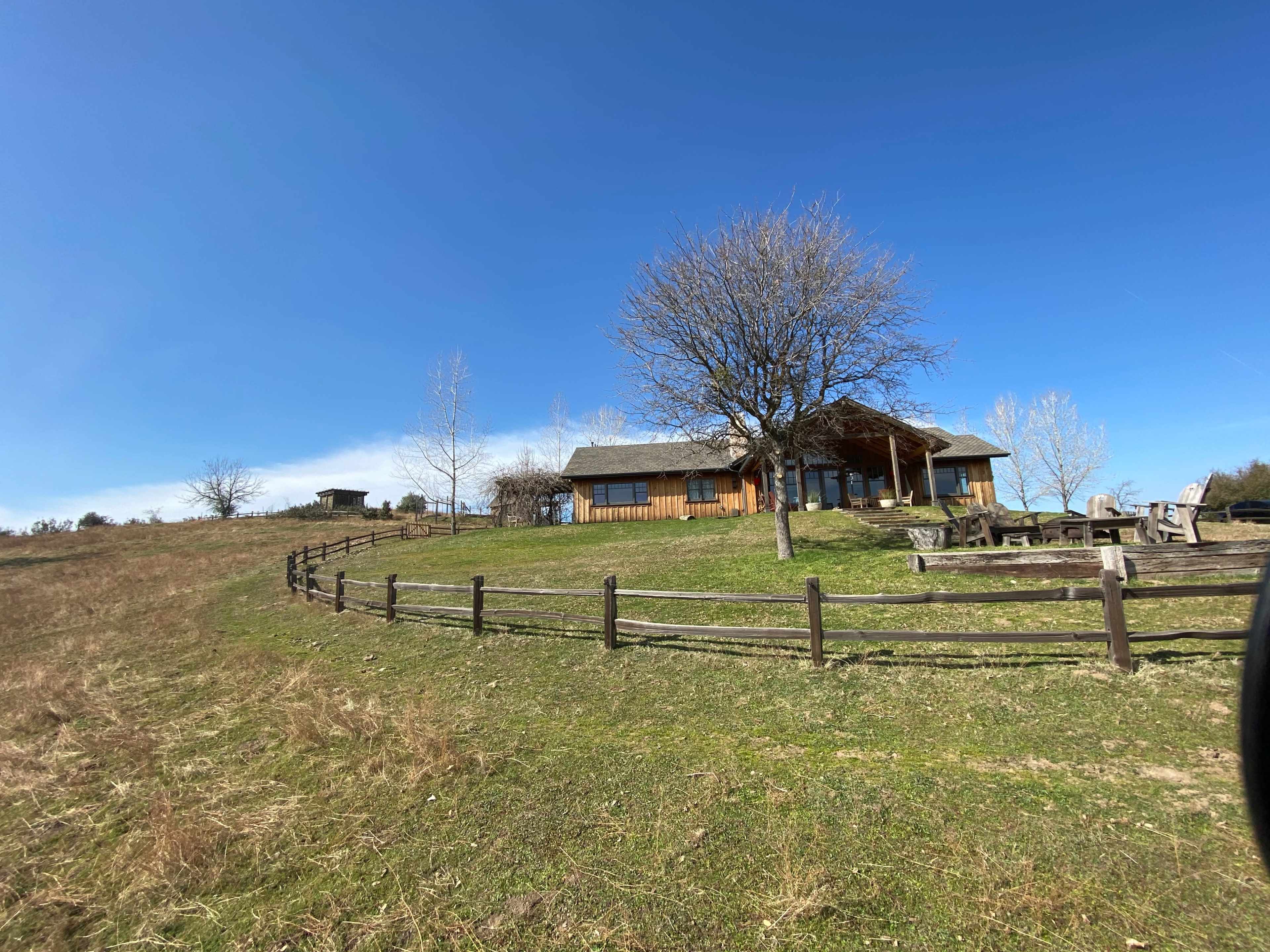 A wooden house sits on a hillside surrounded by a fence and sparse trees under a clear blue sky.