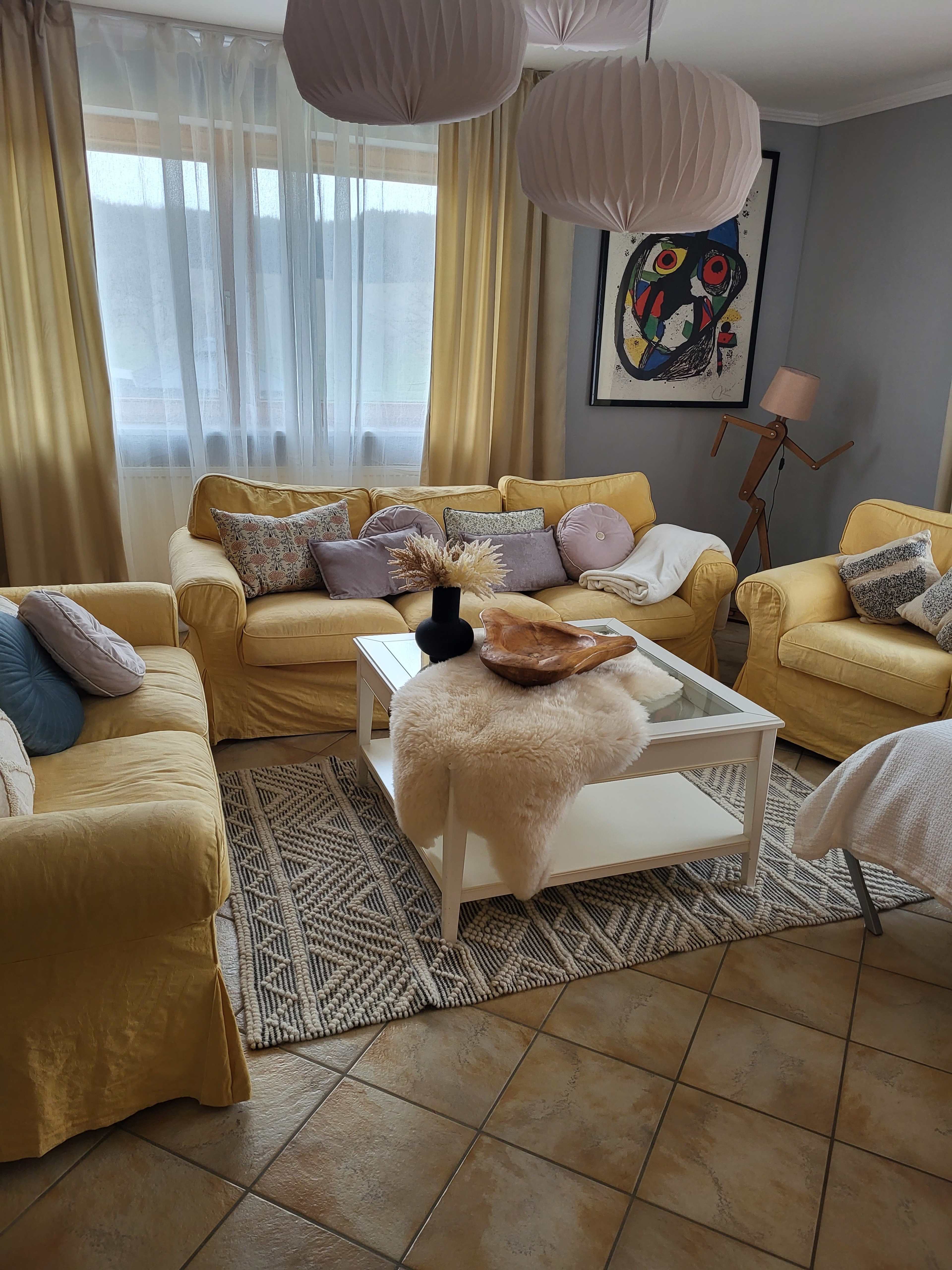 A cozy living room with yellow sofas, a white coffee table, decorative pillows, and a patterned rug under soft lighting.