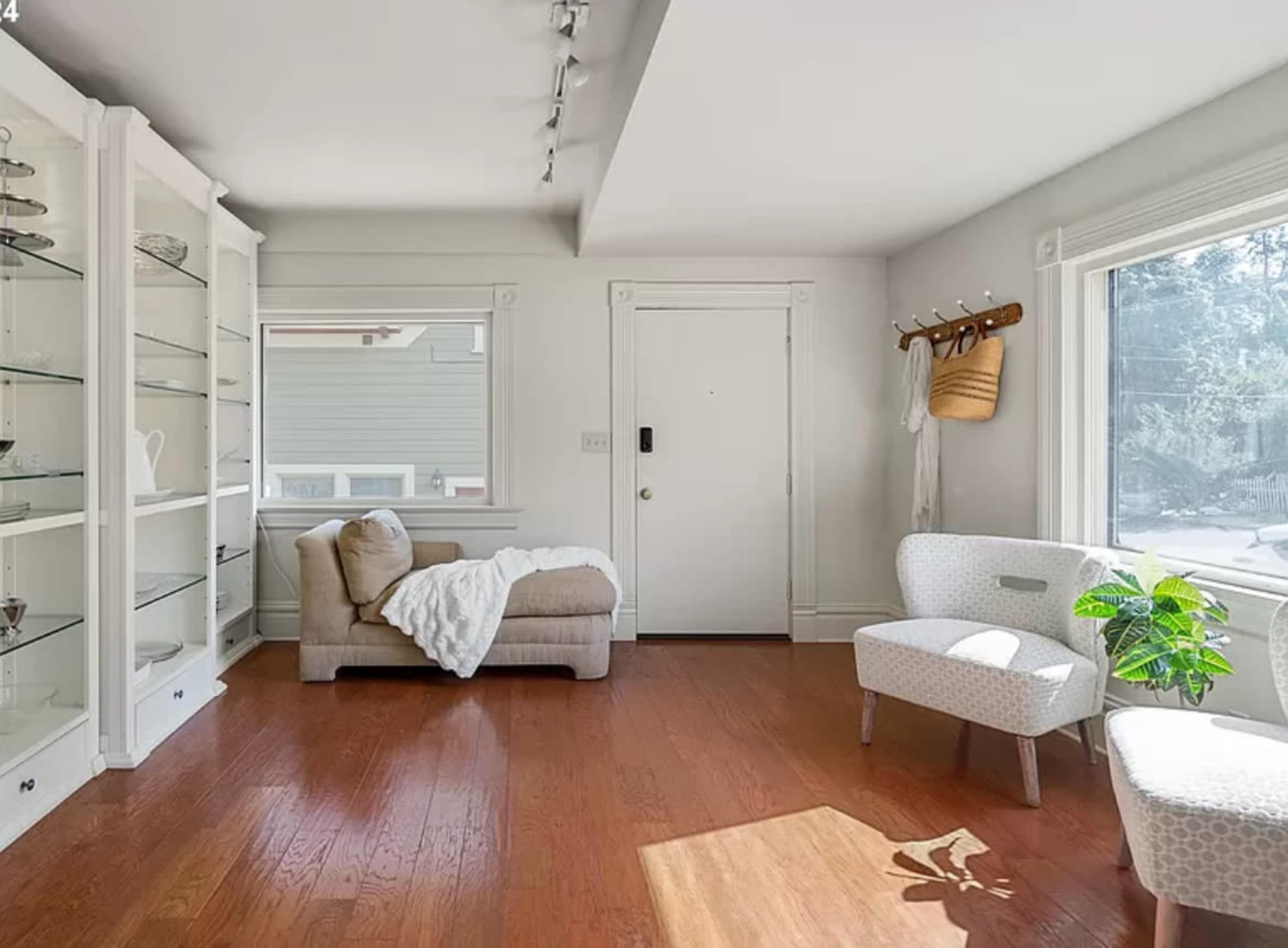 A bright room with a light-colored couch draped in a blanket, a glass display cabinet, and a small chair beside a window with a plant on the floor.