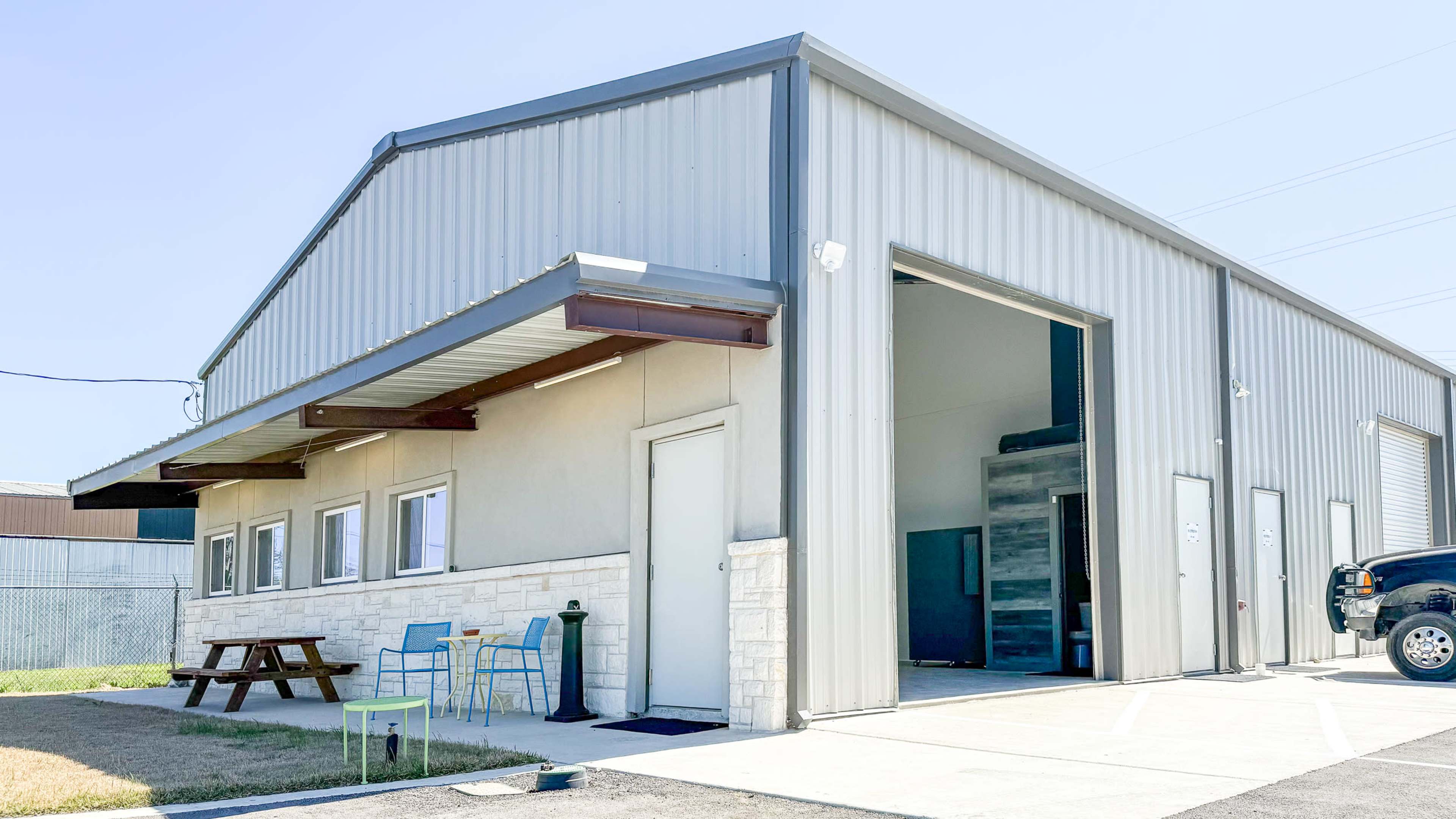 The image shows a metal commercial building with a large opening, a picnic table and chairs outside, and additional doors on the side.