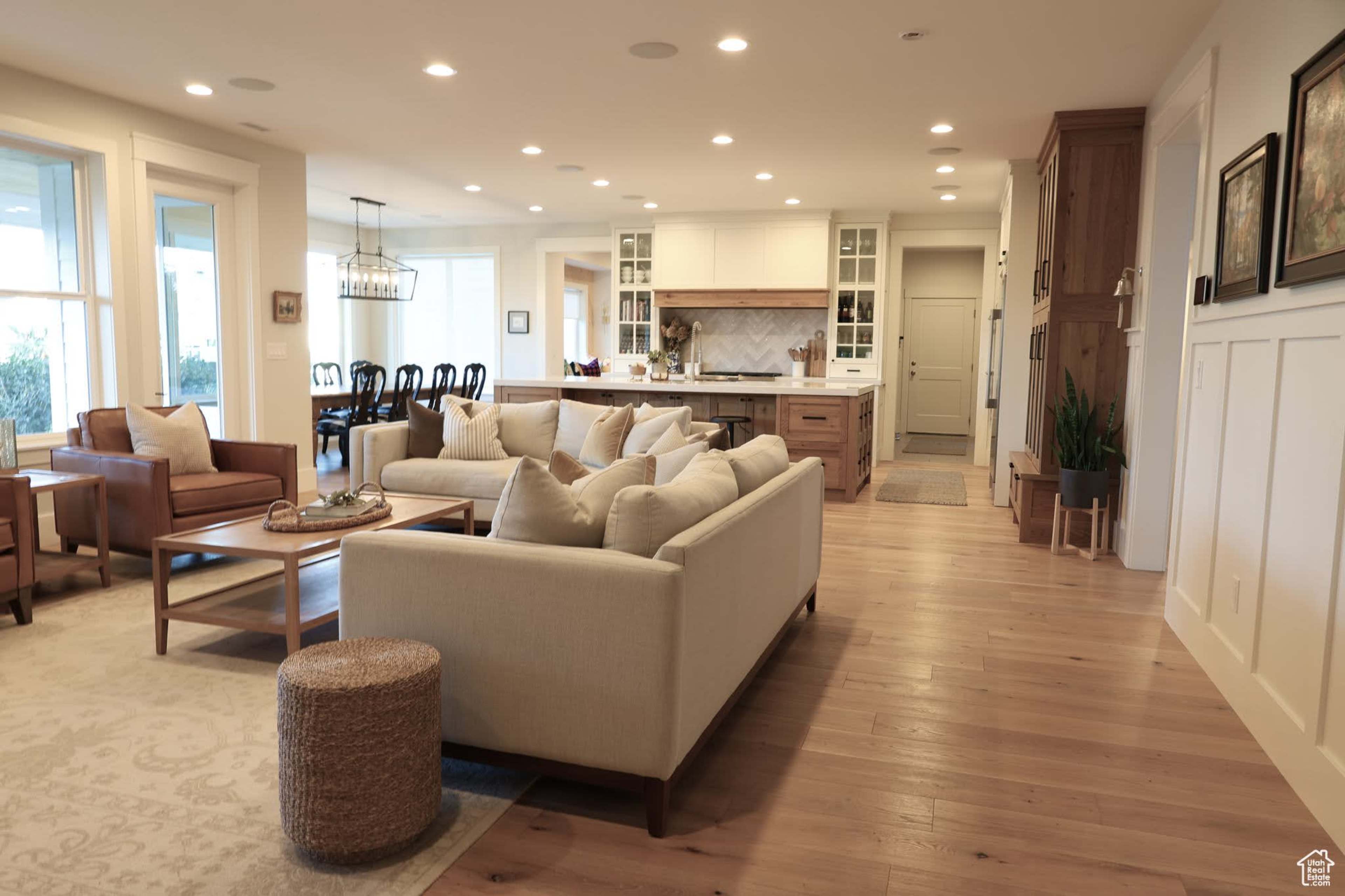 The image shows a modern open-concept living space featuring a large light-colored sofa, wooden furniture, and a kitchen visible in the background.