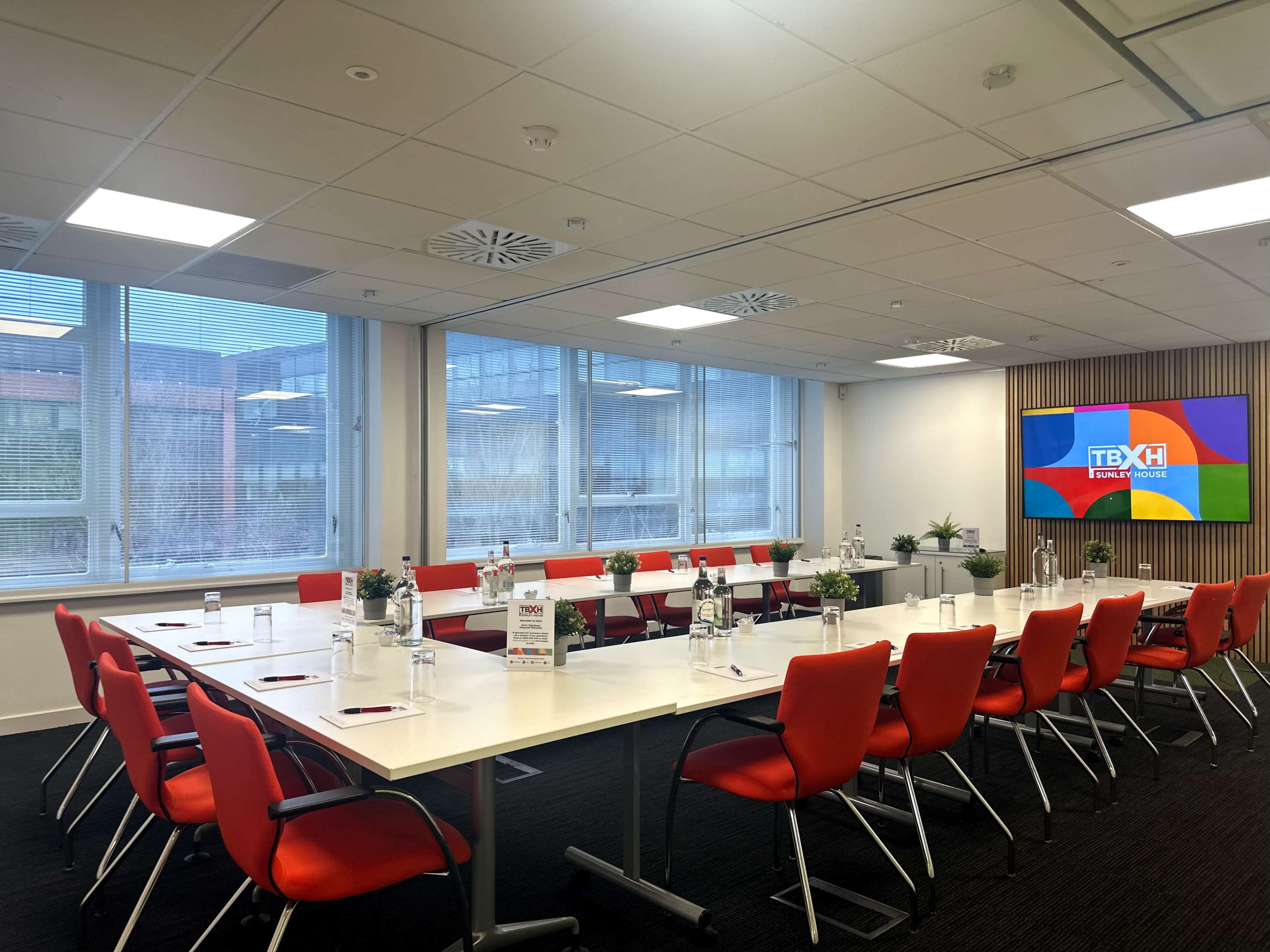 A modern conference room features a long table with red chairs, glass windows, and a large screen displaying a logo.