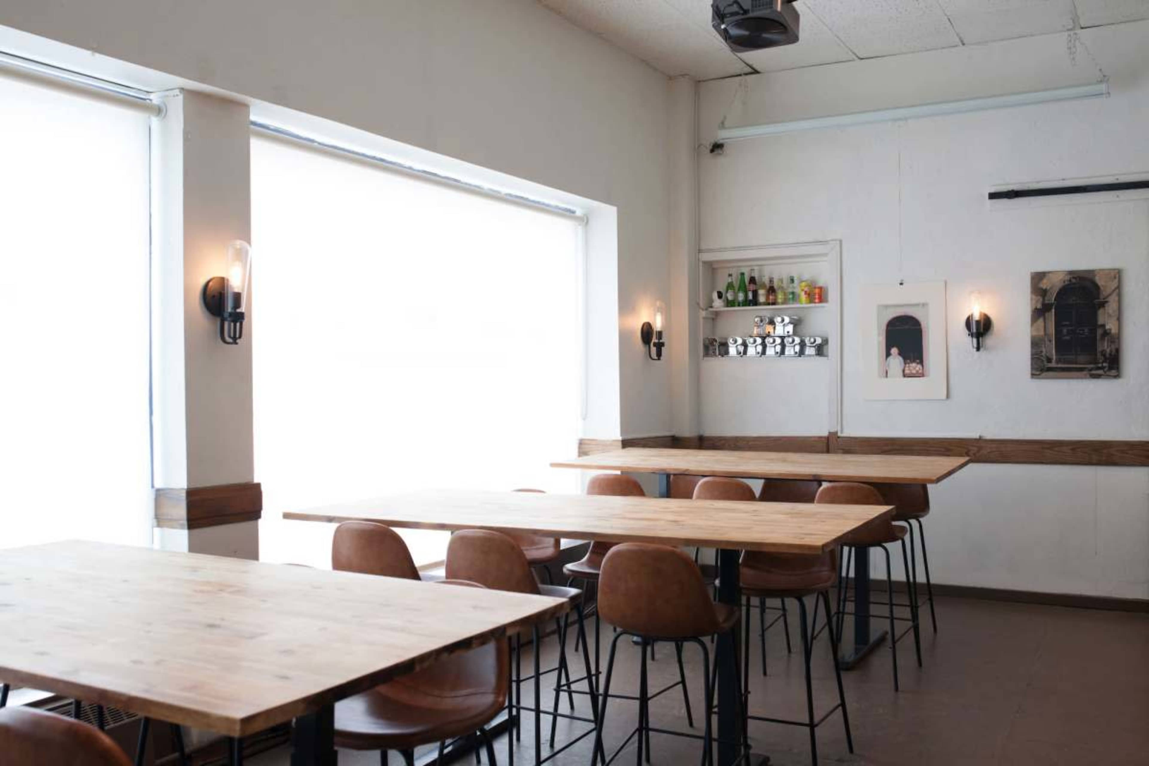A minimalist café interior features wooden tables and brown chairs, with large windows letting in natural light and shelves displaying bottles in the background.