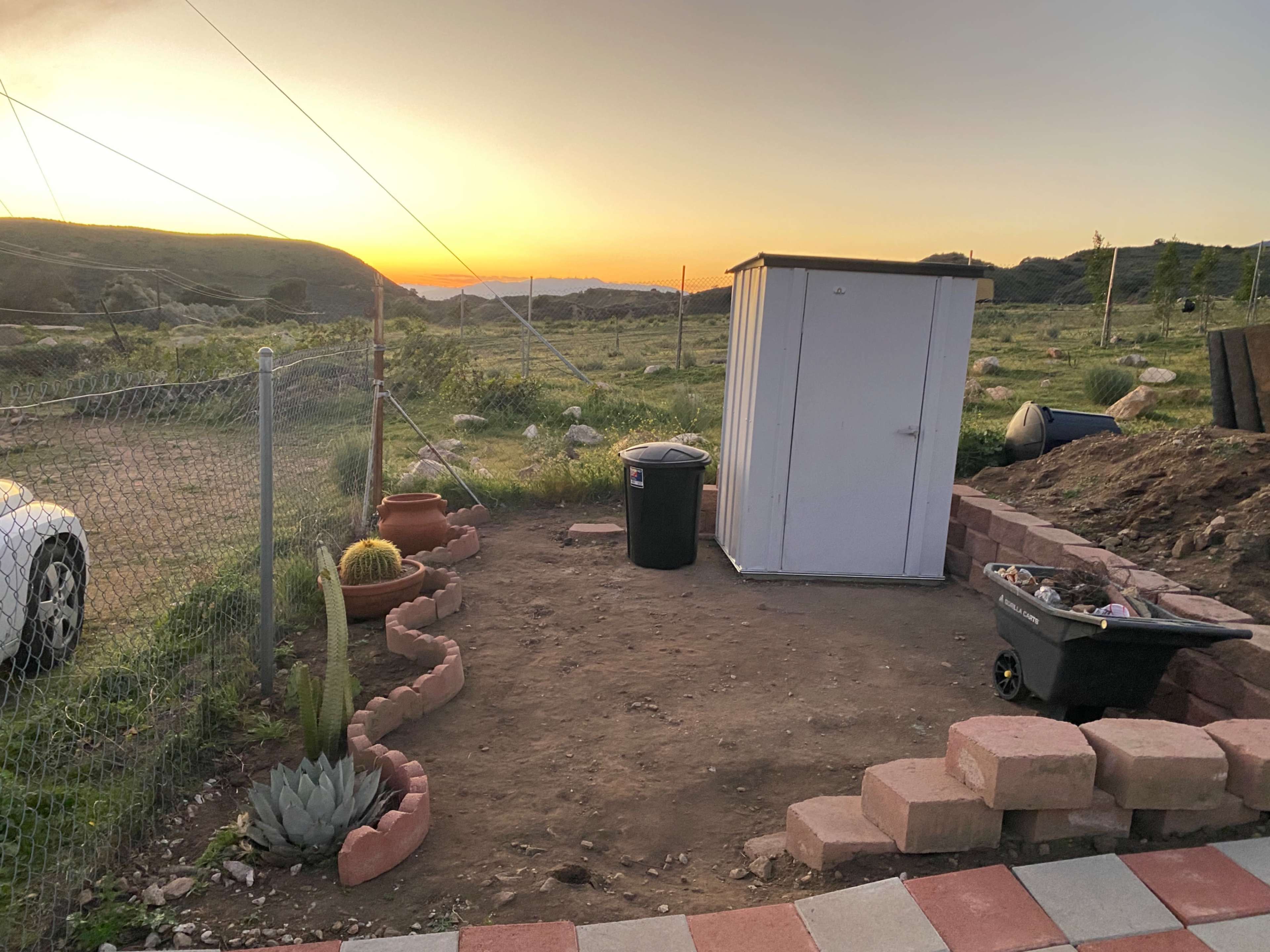 The image shows a small outdoor storage shed next to a garden area with landscaping, a wheelbarrow, and a cactus, set against a sunset backdrop.