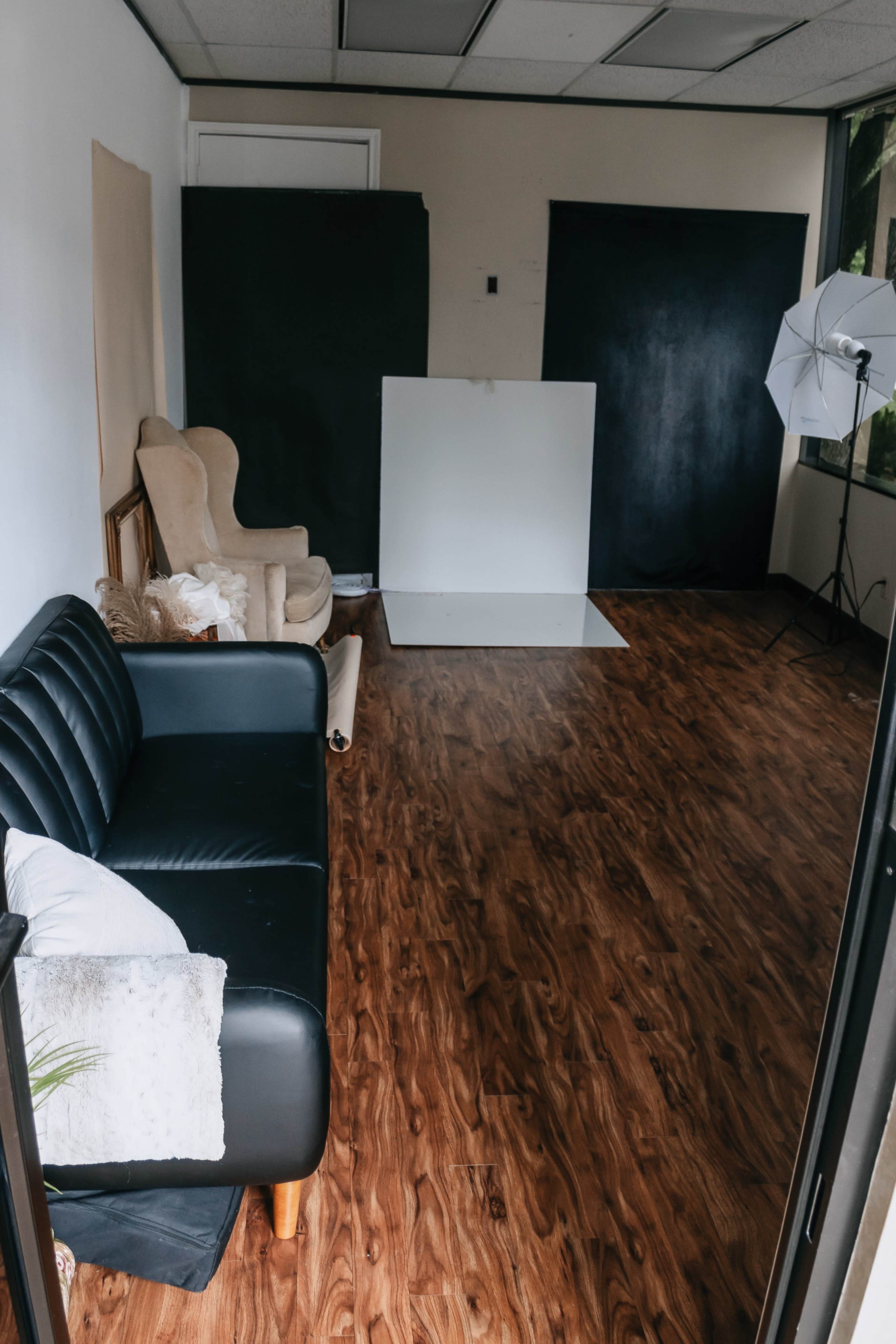 A spacious, empty room with a black leather couch, an armchair, and a photography backdrop set up against the wall.