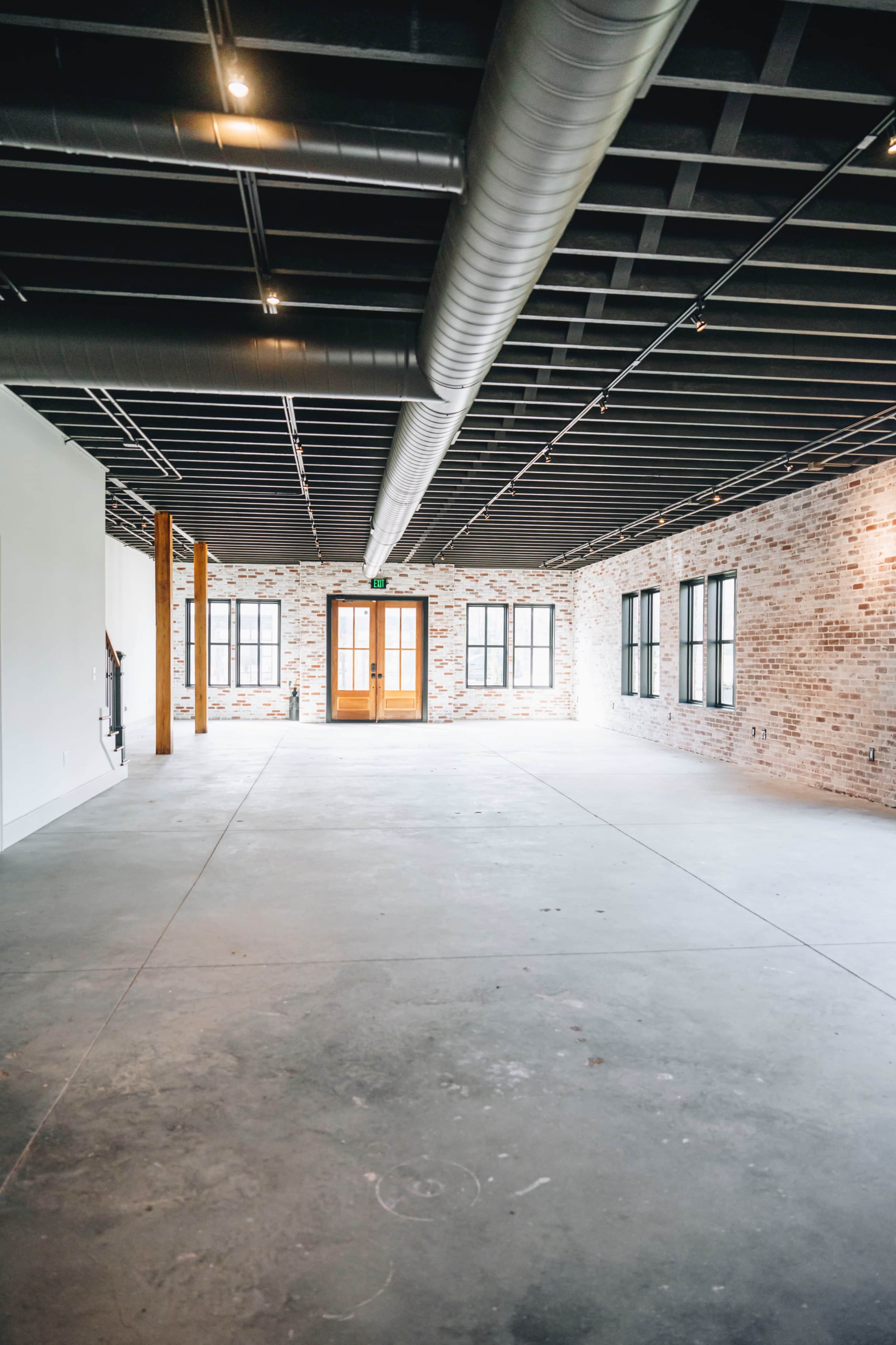 A large, empty room with exposed brick walls, concrete flooring, and large windows, featuring a set of double doors at one end.