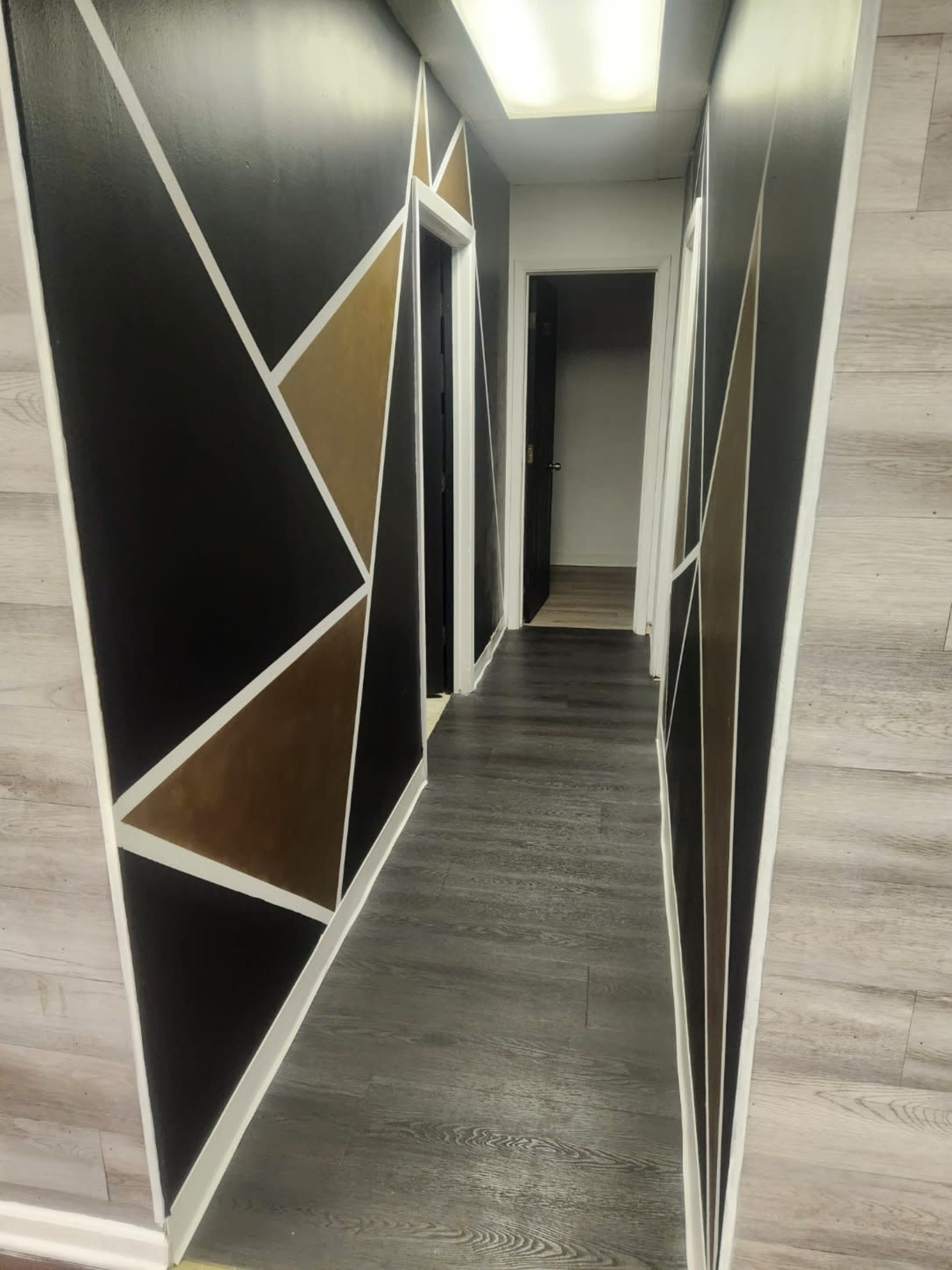 The image shows a narrow hallway with dark walls featuring geometric gold and white designs, leading to open doorways at the end.