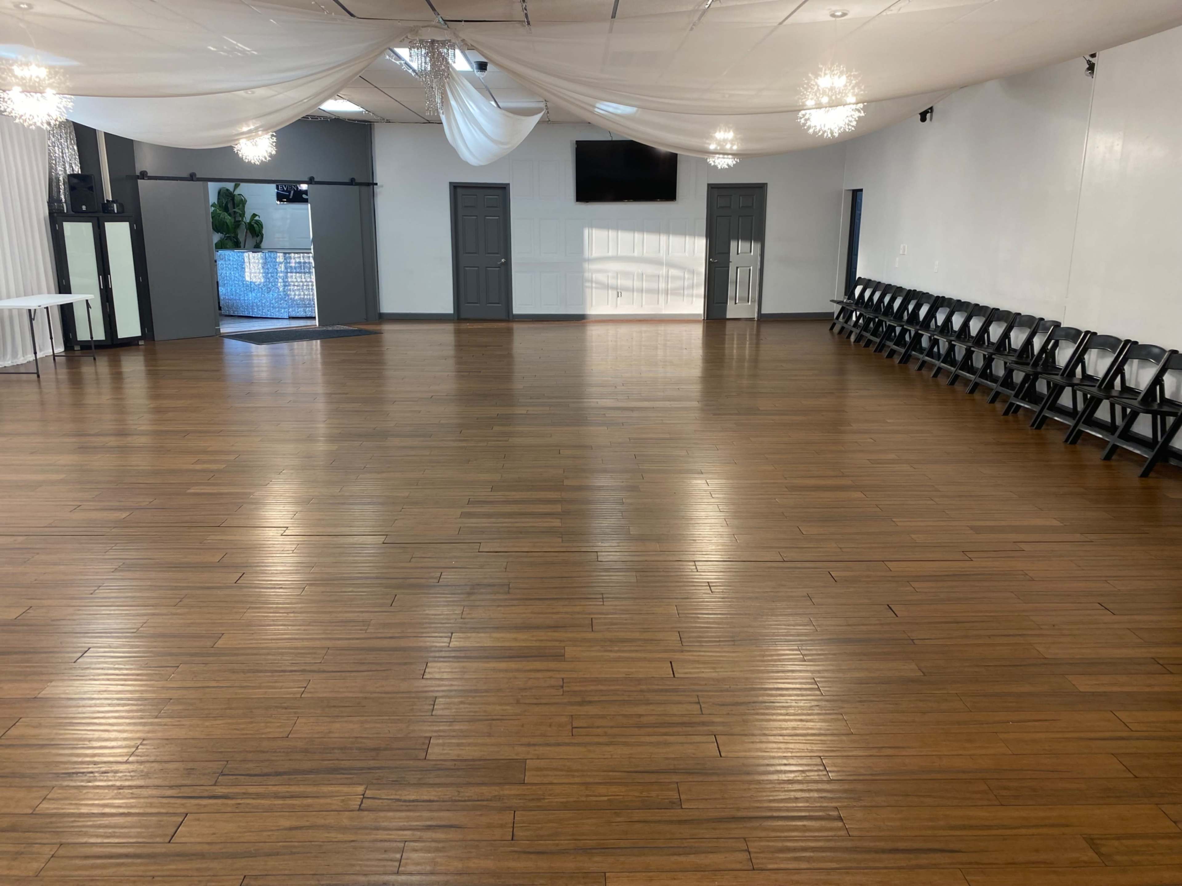 A spacious event hall with a polished wooden floor, white draped ceiling, and rows of black folding chairs against one wall.