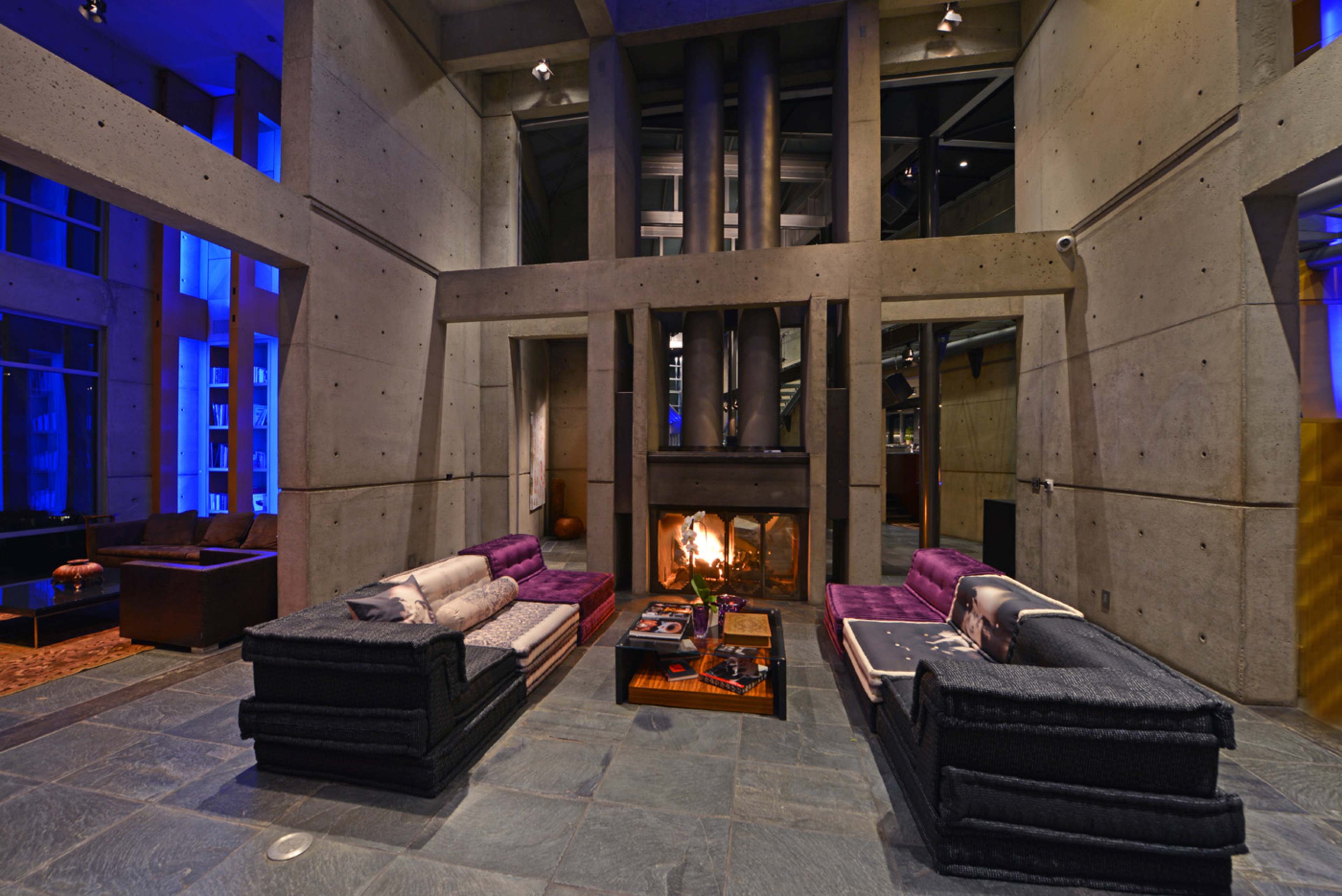 The image shows a modern living room featuring large concrete walls, two sofas facing a central coffee table, and a fireplace with flames visible.