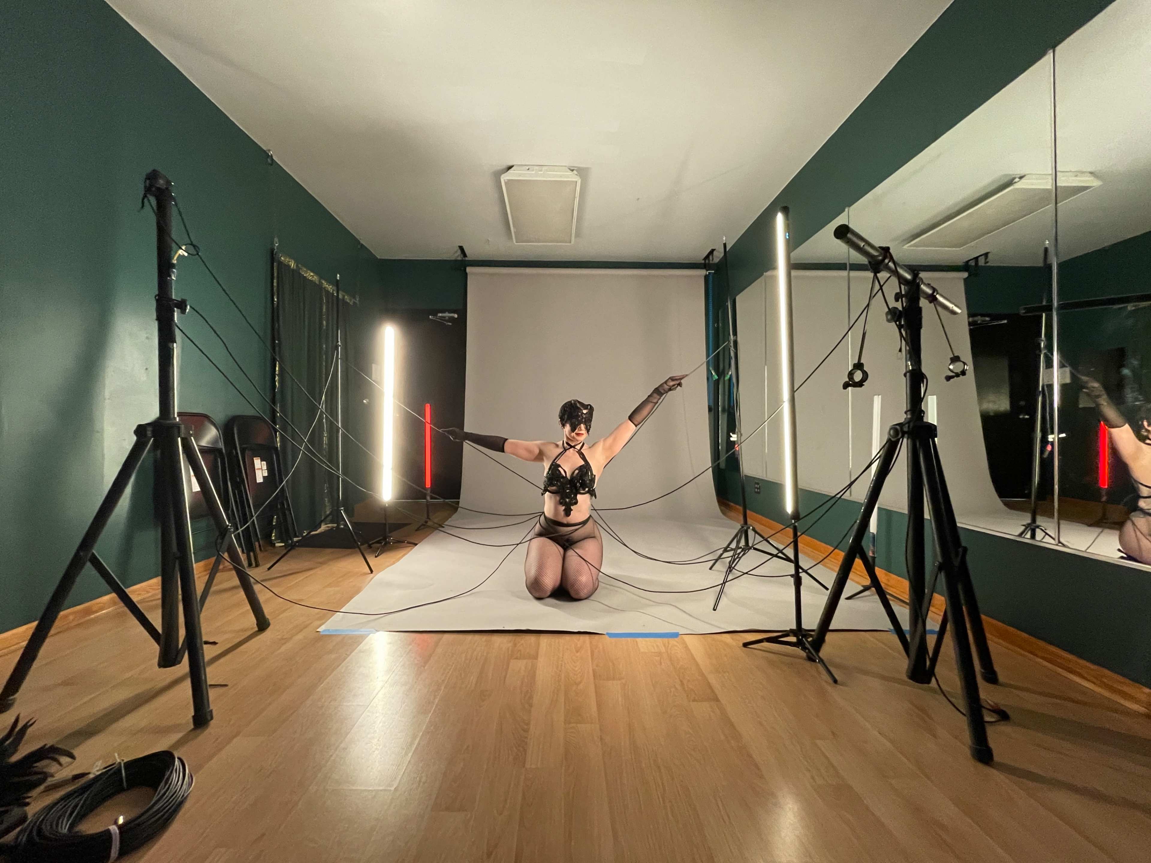 A performer dressed in a black outfit poses on a gray backdrop surrounded by studio equipment and lighting.