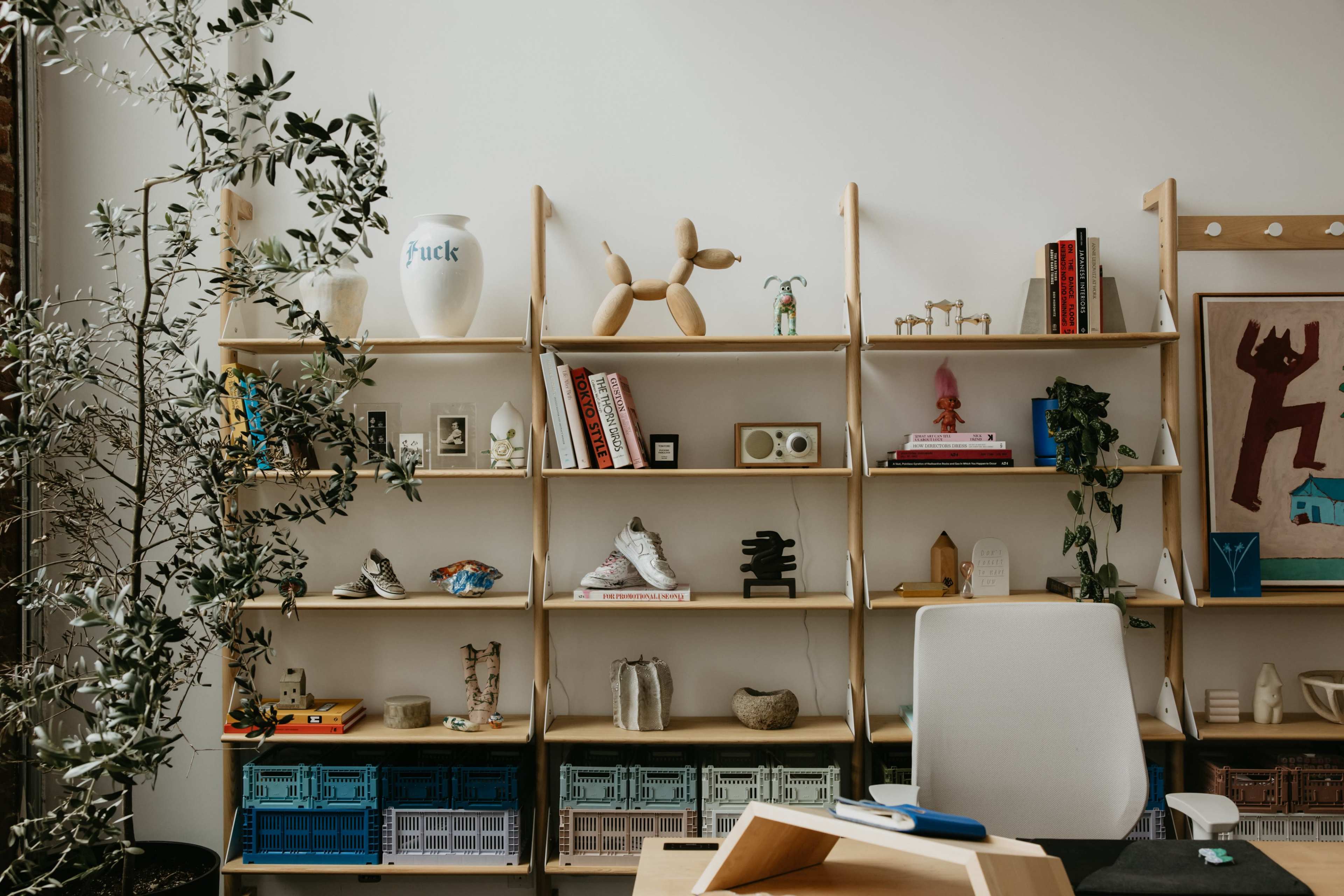 The image shows a wooden shelving unit filled with various decorative items, books, and a potted plant, alongside a white office chair and a wooden desk.