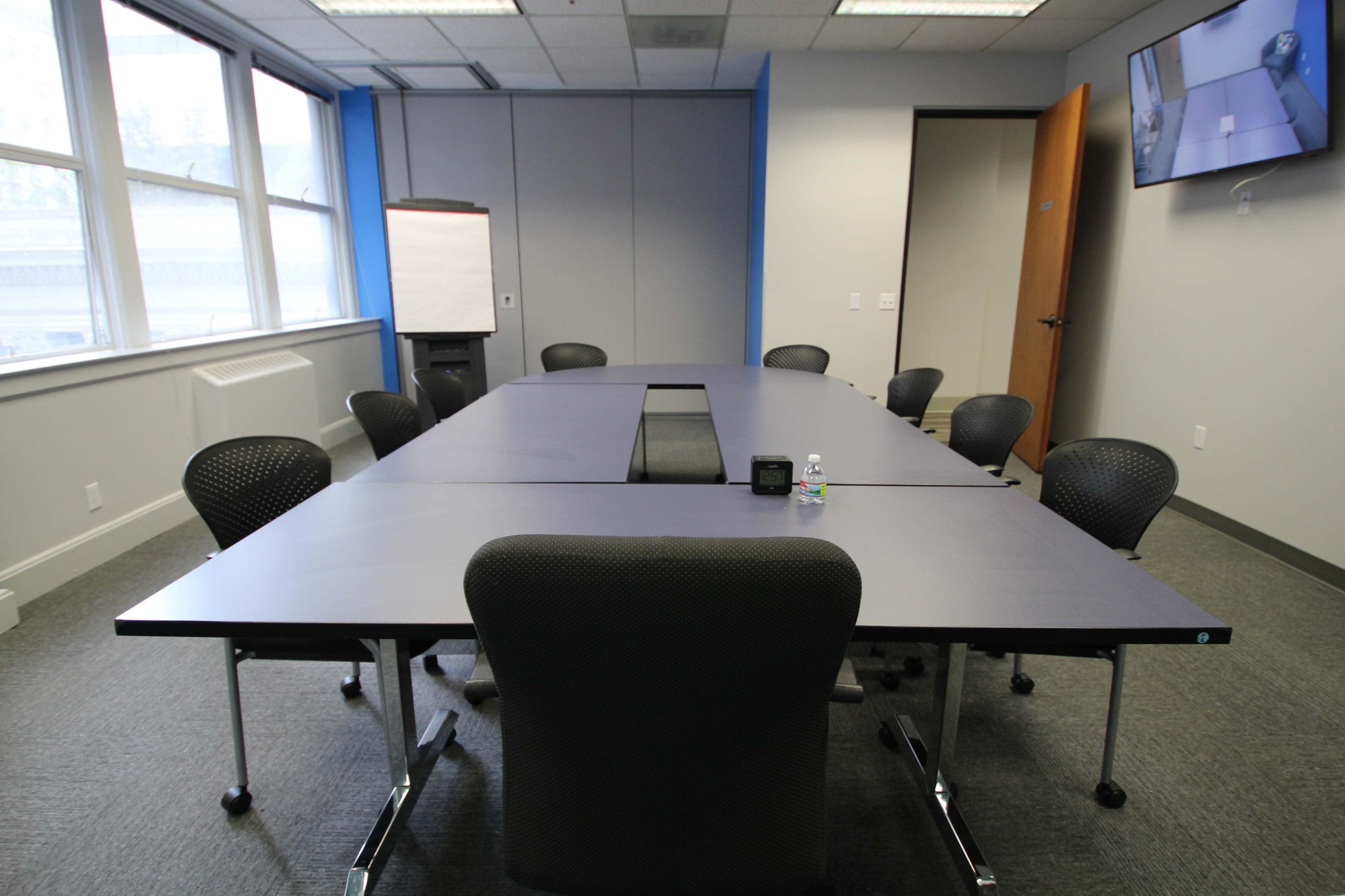 A large conference room features a long table surrounded by black chairs, with a whiteboard and a monitor on the walls.