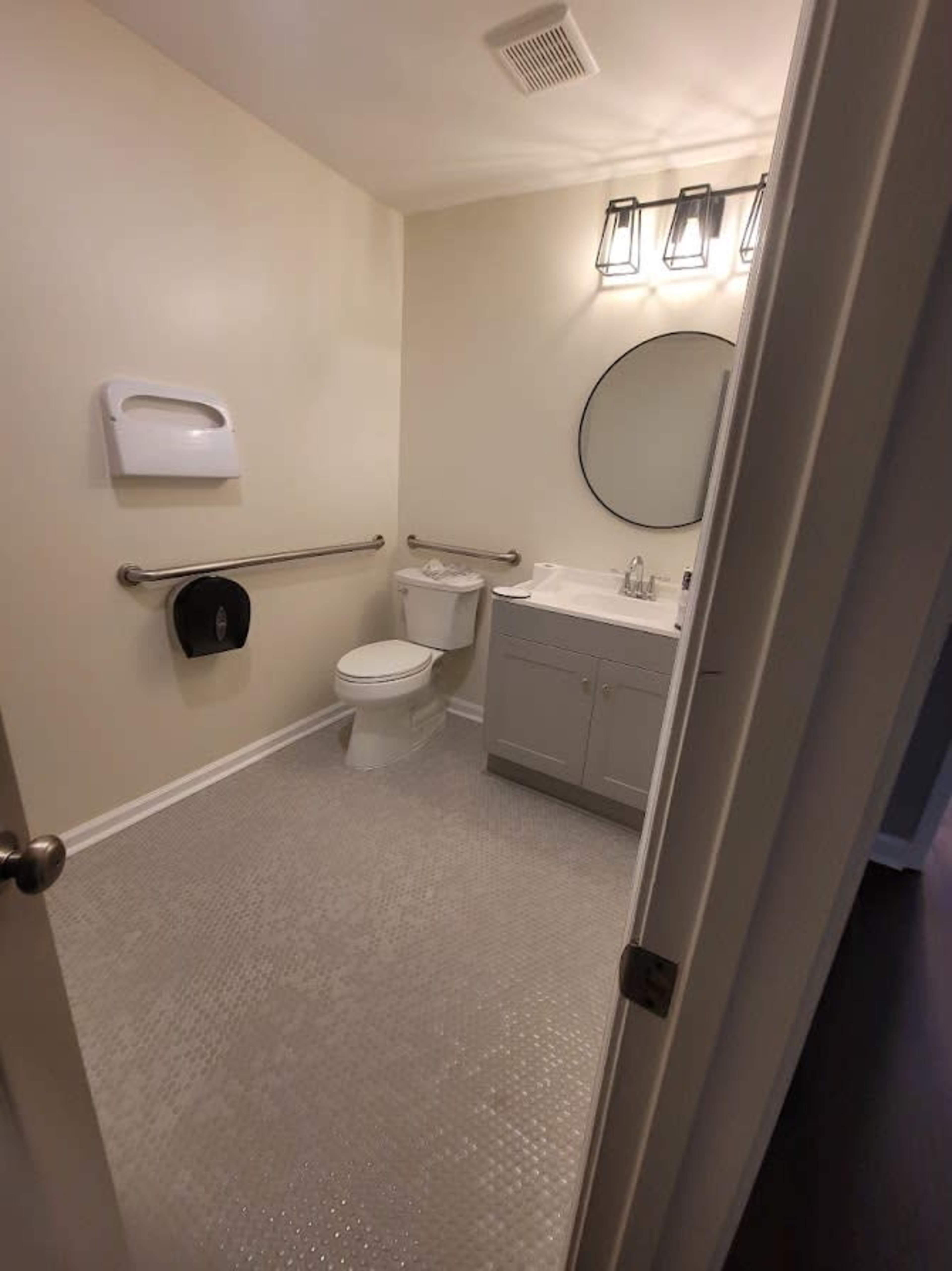 A clean bathroom features a toilet, a sink with a vanity, a mirror, and grab bars, all situated on a tiled floor.