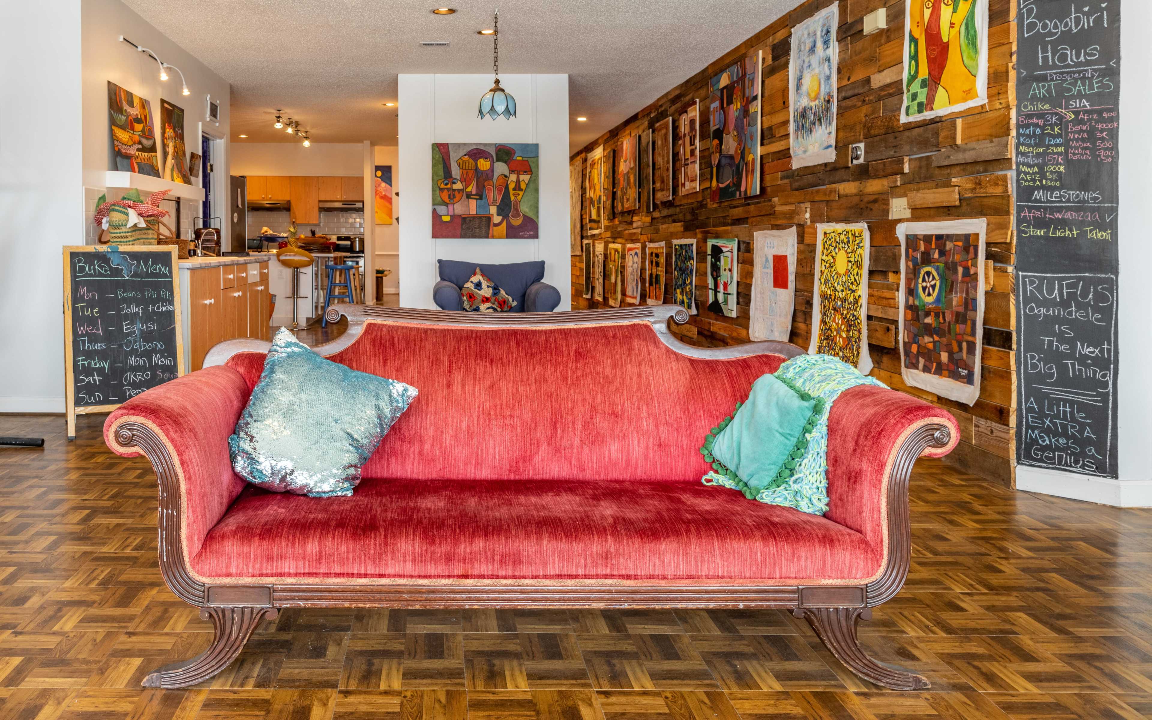 A red vintage couch with decorative pillows is positioned in a room featuring wooden walls adorned with colorful artwork and chalkboard menus.