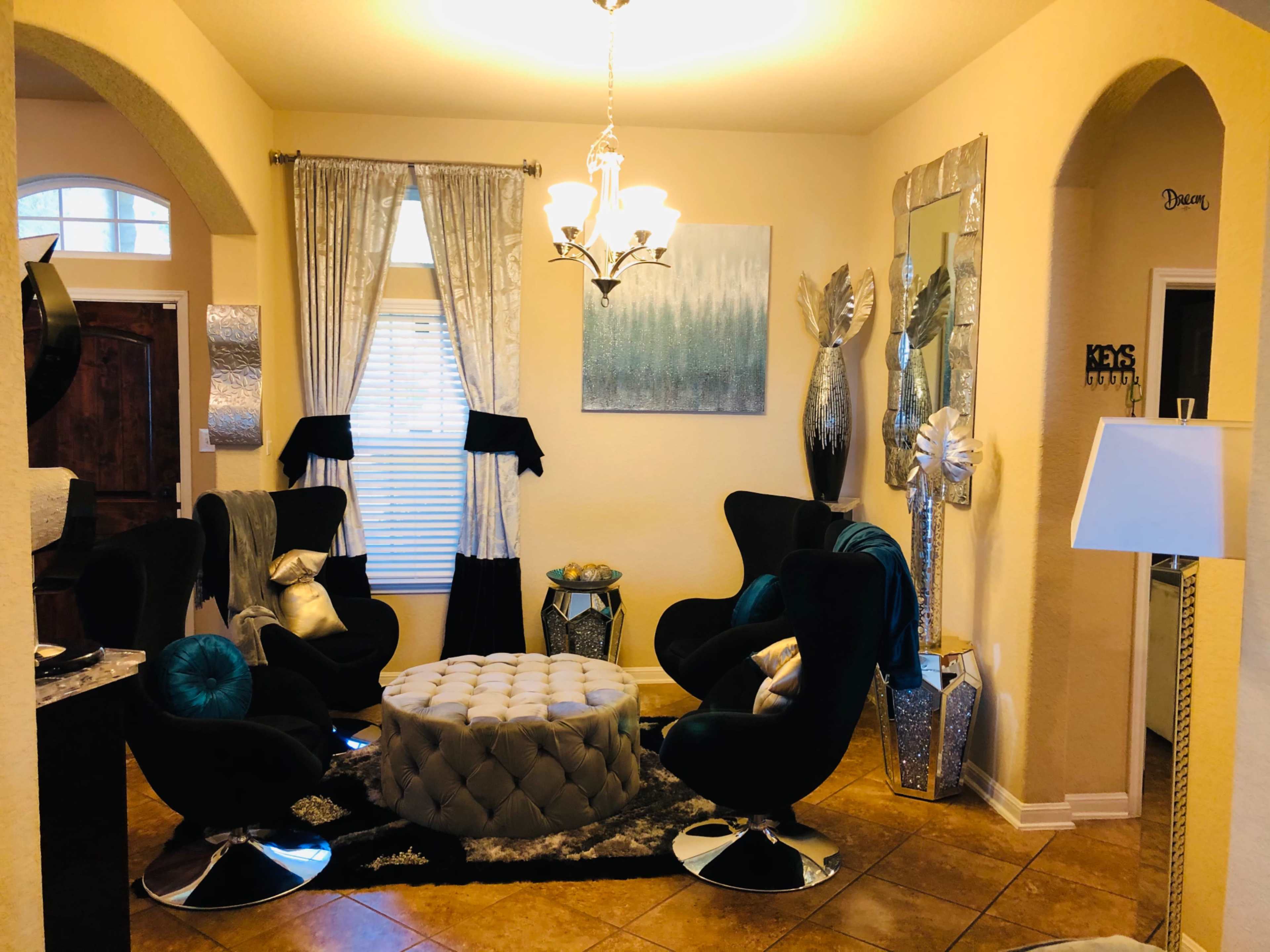 The room features a round tufted coffee table surrounded by four black and teal chairs, with elegant decor including a chandelier and decorative wall mirrors.