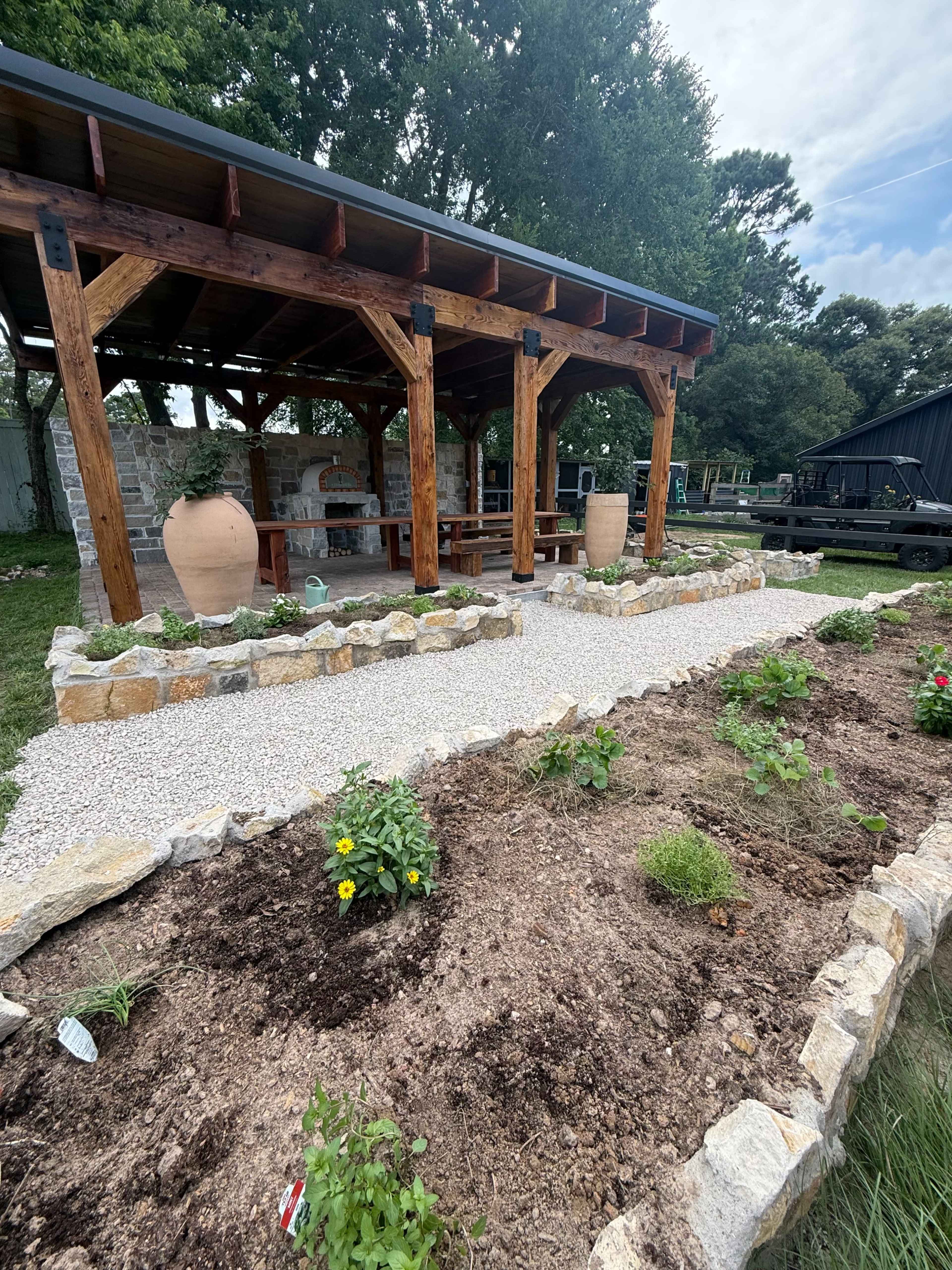 The image shows a wooden gazebo structure with a stone path and a garden featuring various plants in front of it.