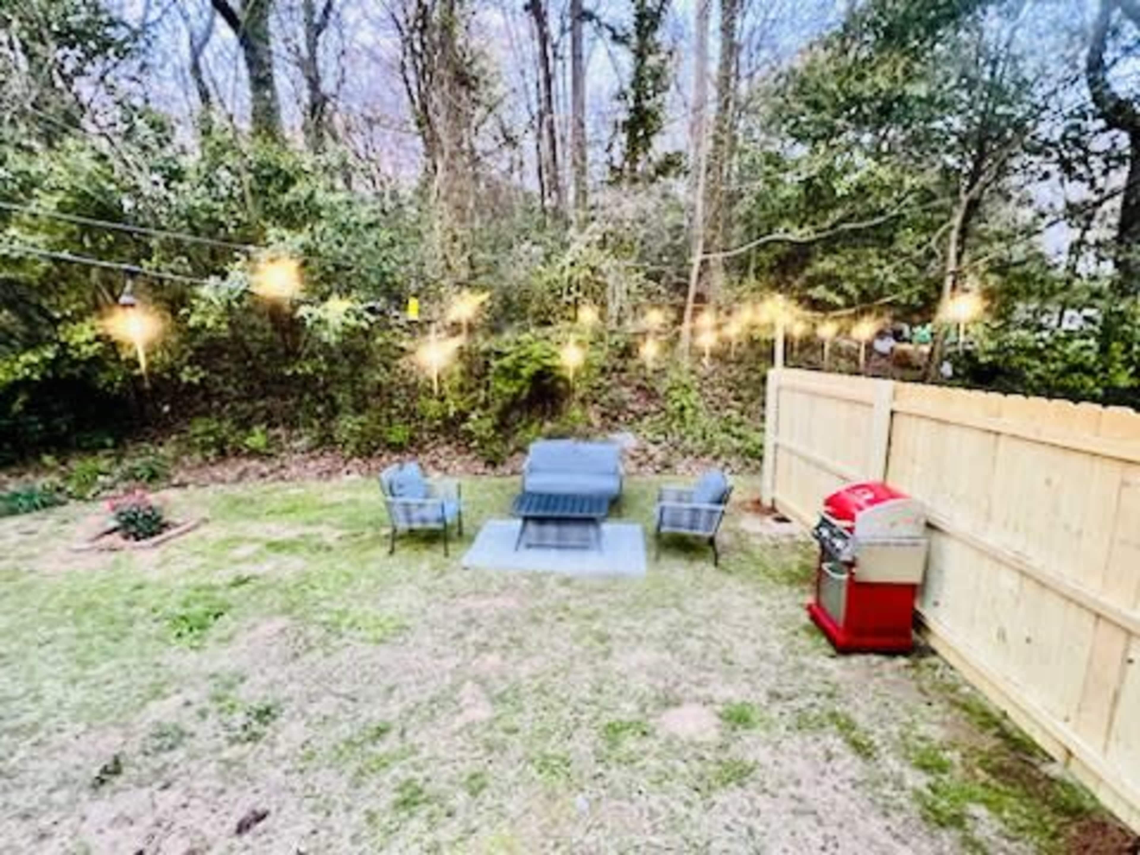 A backyard setup with four chairs arranged around a fire pit, string lights overhead, and a red grill placed against a wooden fence.