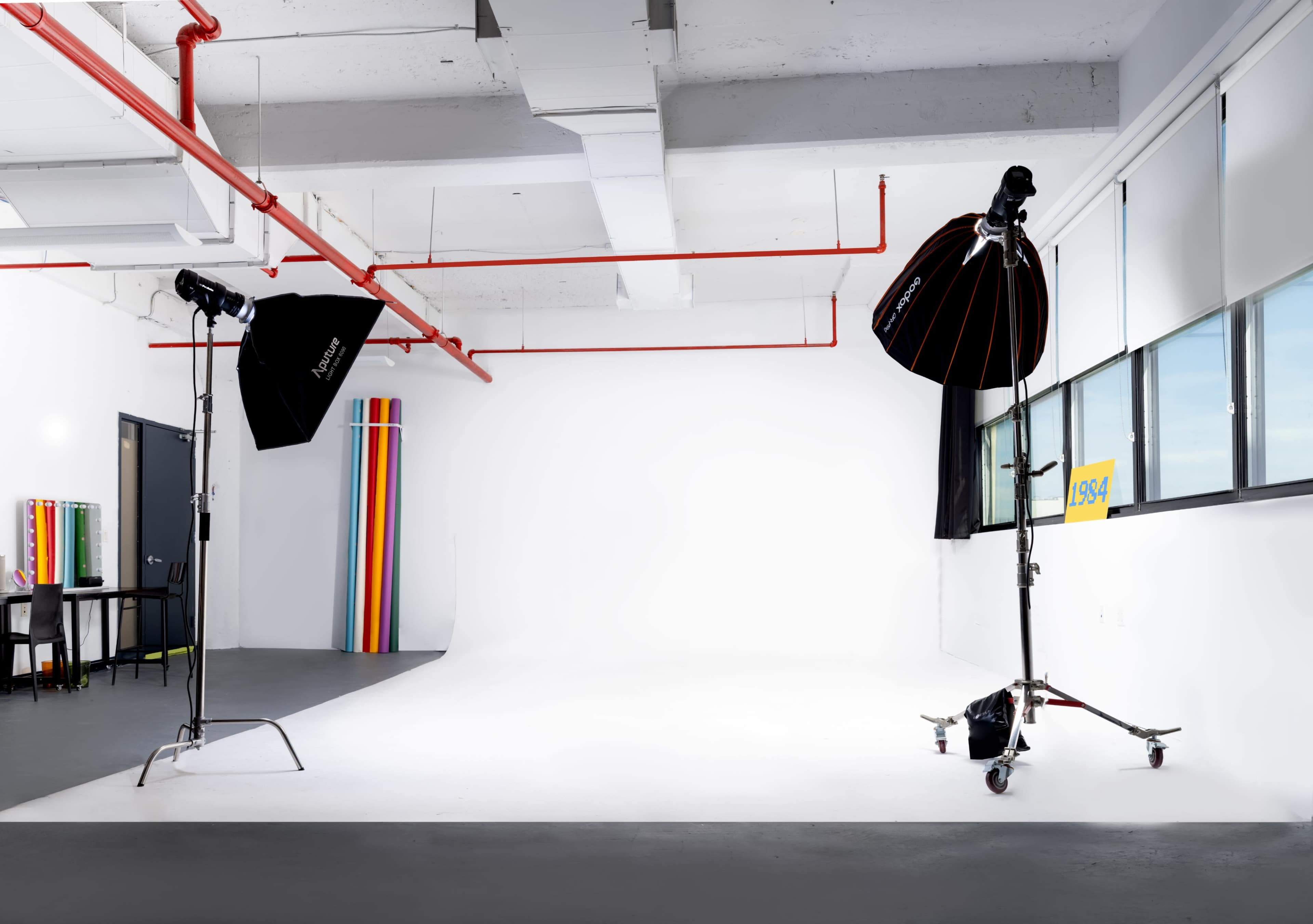 The image shows a photography studio with a white backdrop, lighting equipment, and colored backdrops leaning against a wall.