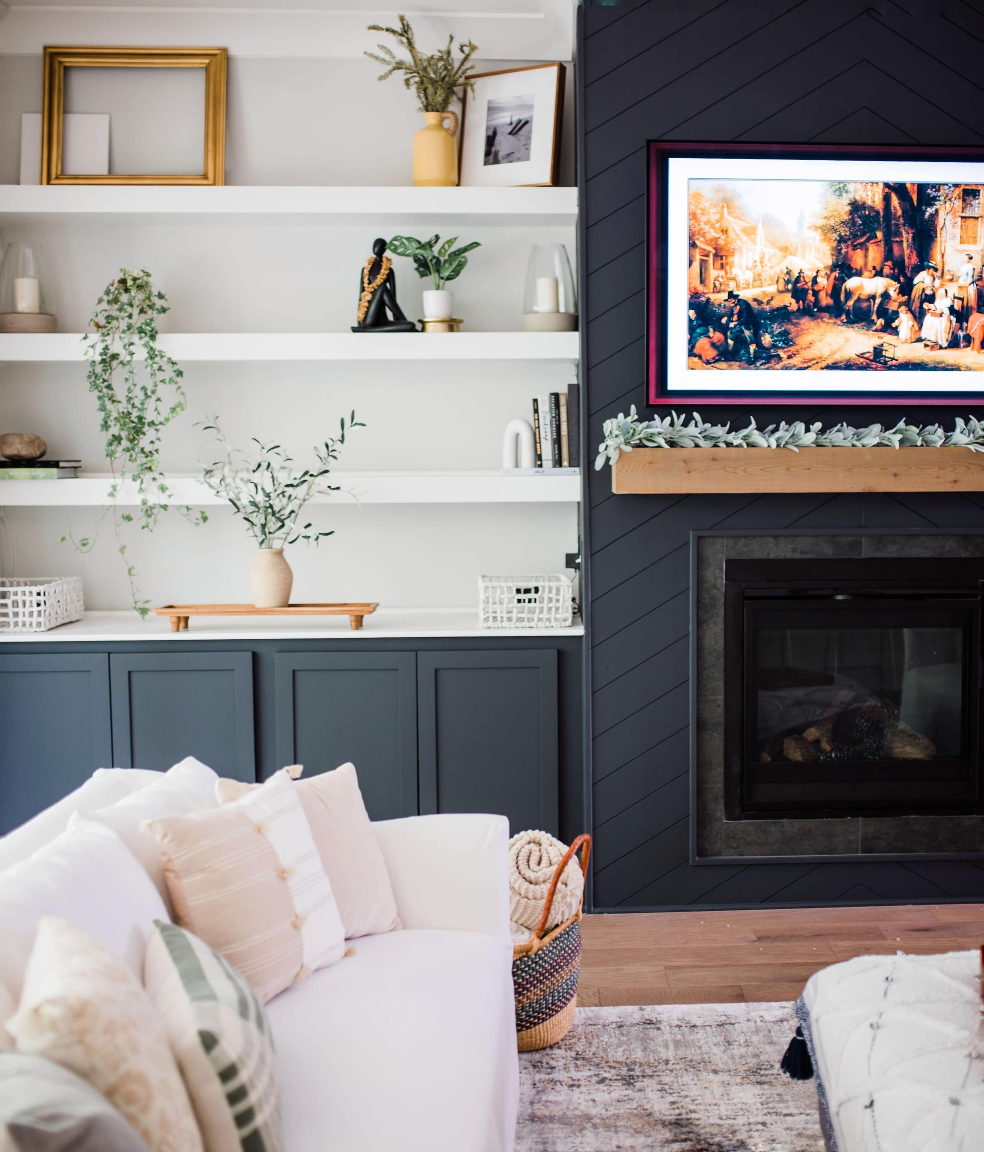 A living room with a white couch, a wooden media console beneath a television, and shelves decorated with plants, vases, and artwork.