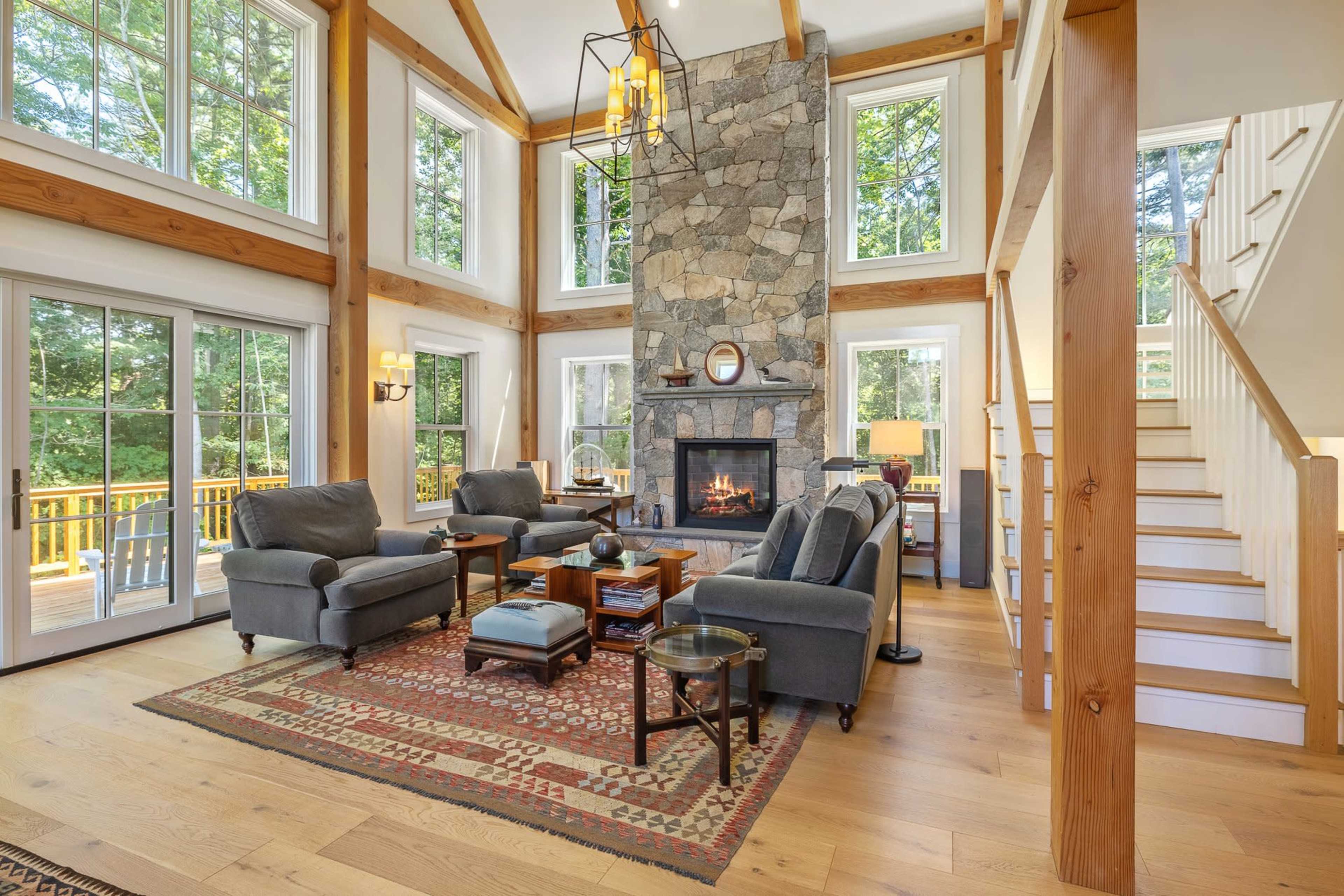 A spacious living room features a stone fireplace, wooden beams, large windows with views of the outdoors, and comfortable seating arrangements.