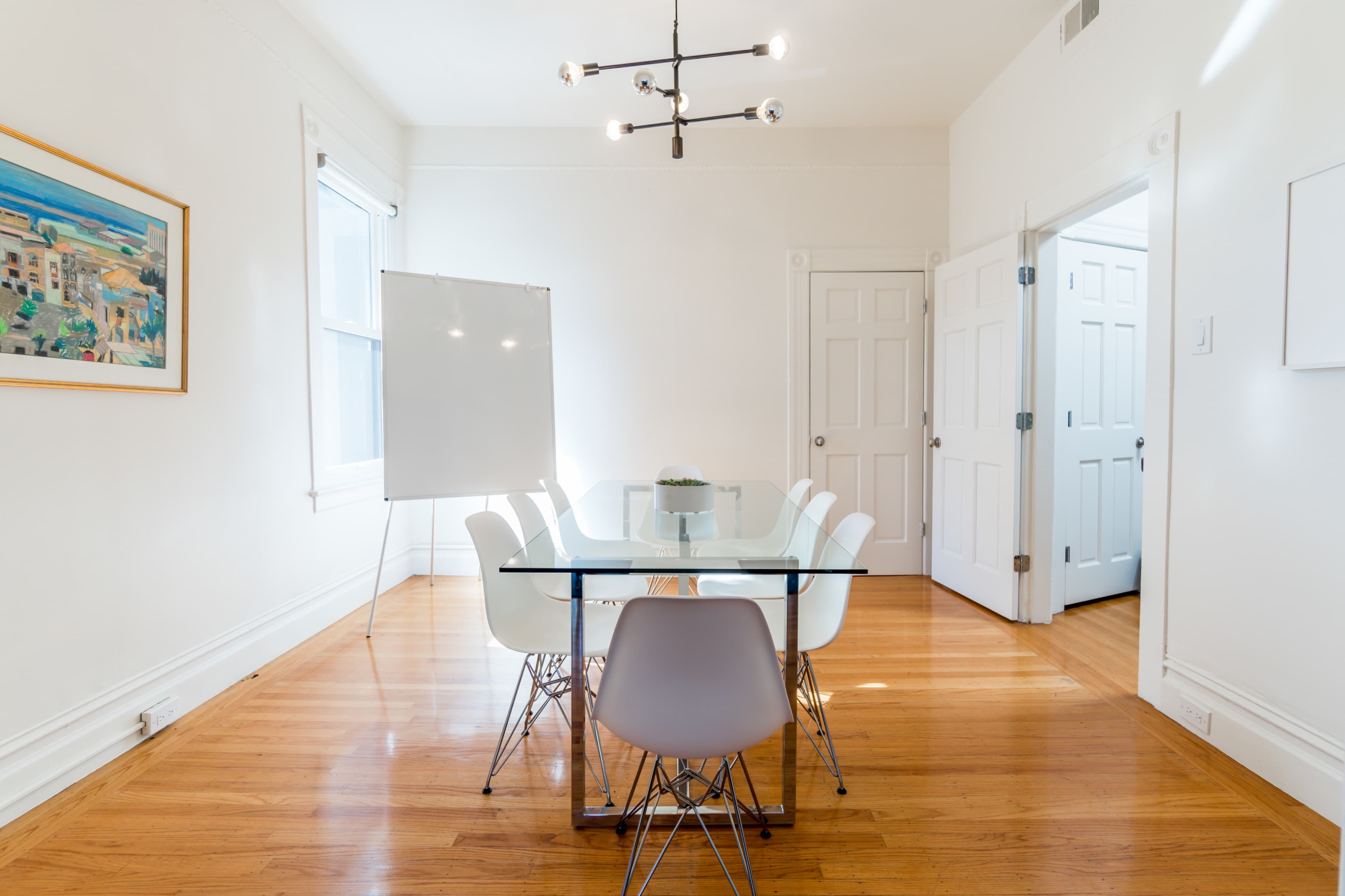 A bright, minimalist conference room features a glass table surrounded by white chairs, with a whiteboard and a door leading to another room.