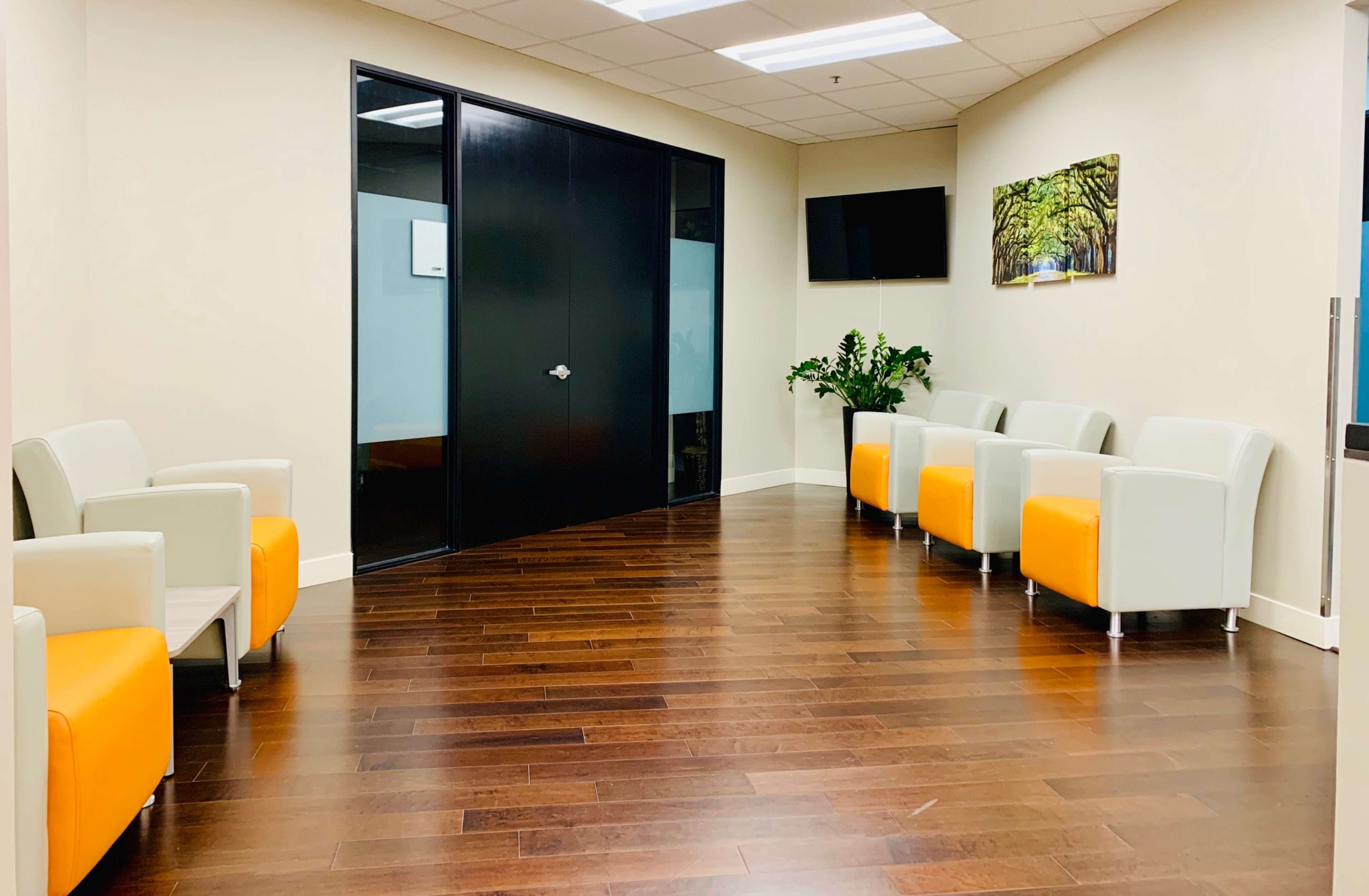 A waiting area with white and orange chairs, wooden flooring, and a glass door leading to an office space.